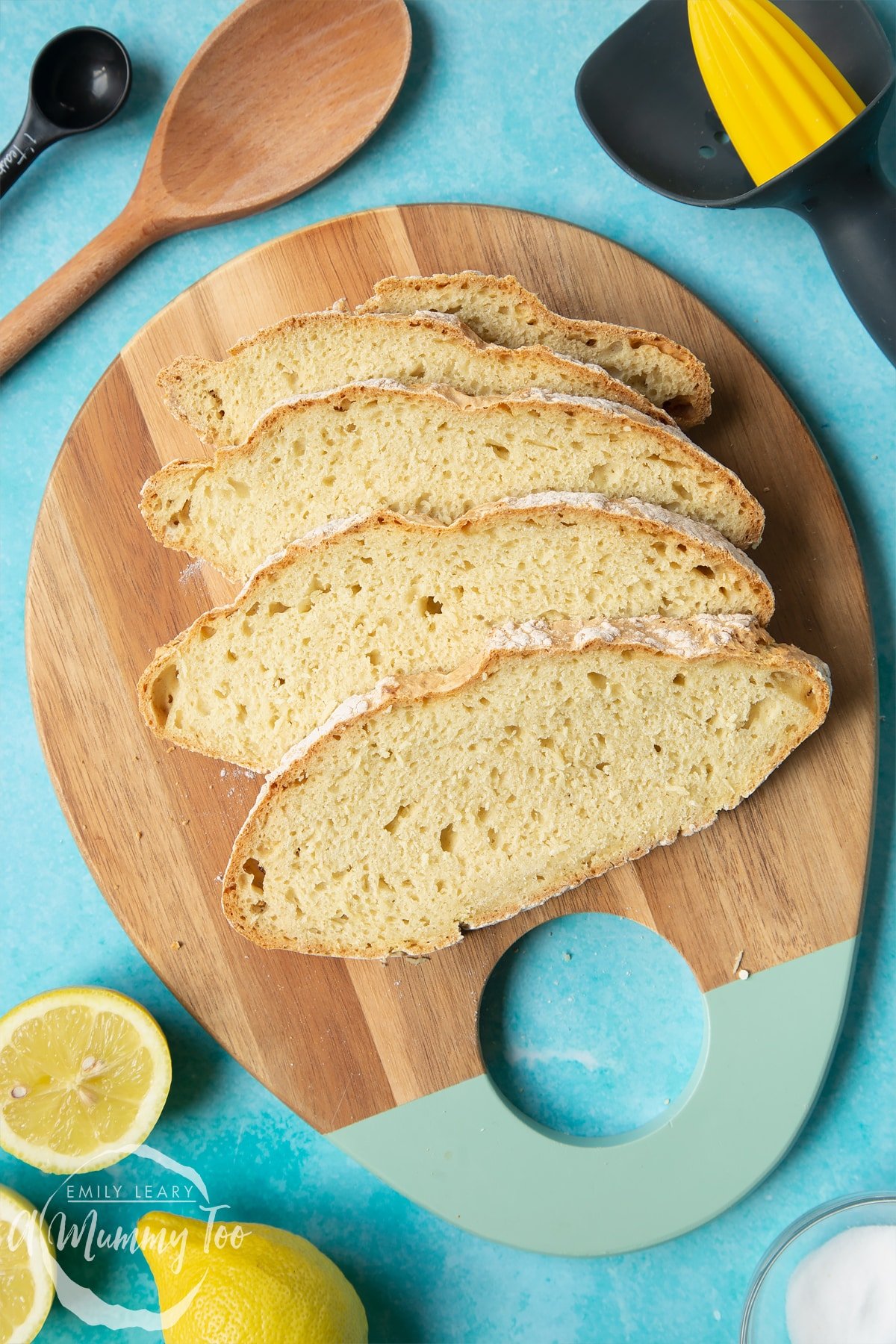 Vegan soda bread slices on a wooden board. Lemons and utensils surround the board.