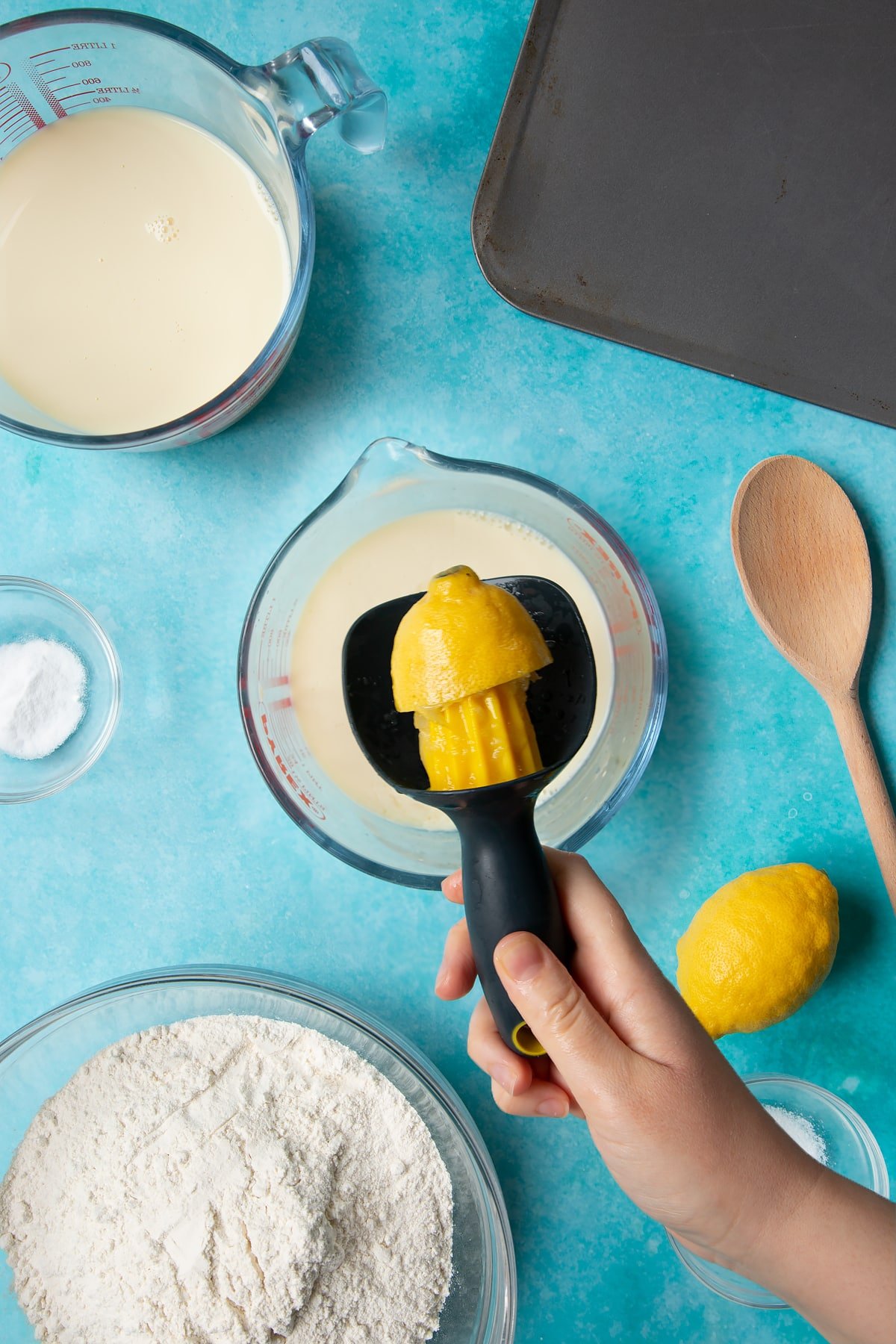 A jug containing plant milk. A hand holds a lemon half on a juicer above the jug. Ingredients to make vegan soda bread surround the jug.