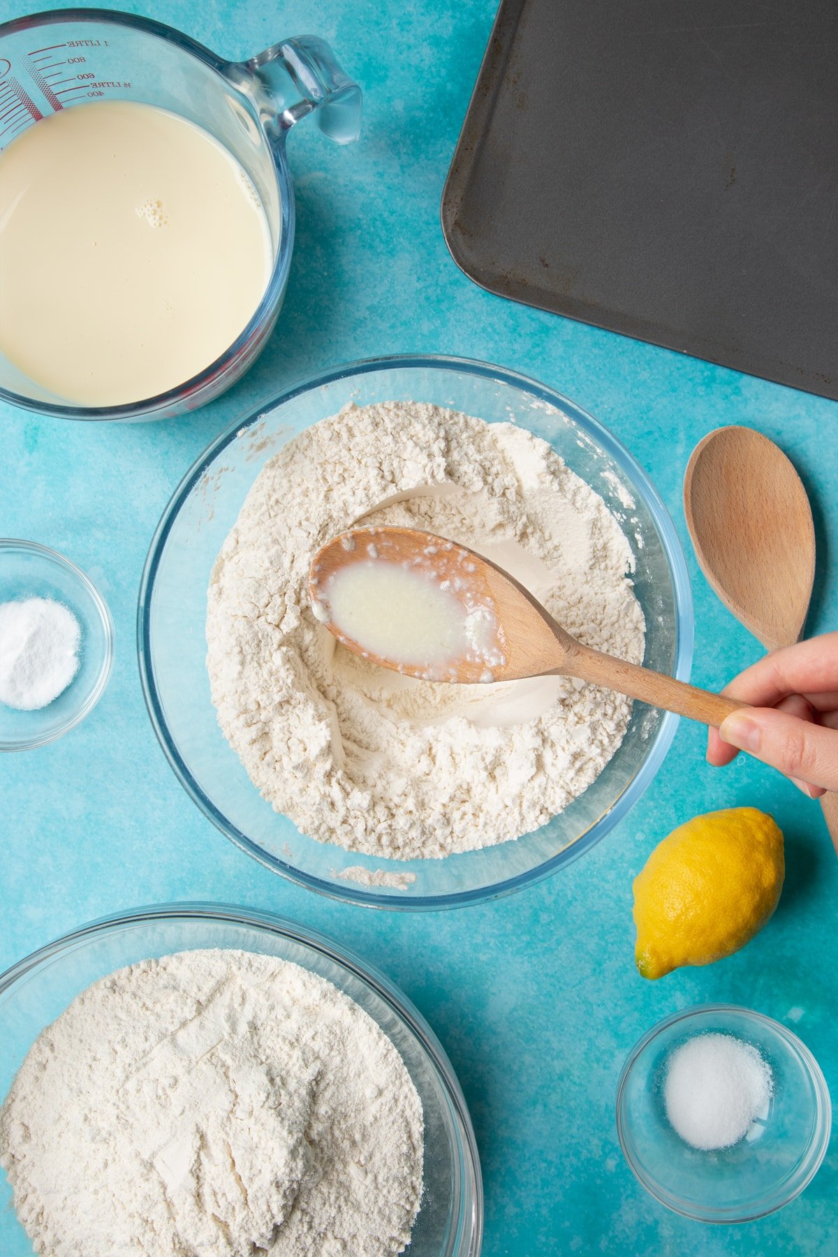 A bowl containing plain flour, salt and bicarbonate of soda. A hand holds a spoon with plant milk on it. Ingredients to make vegan soda bread surround the bowl.