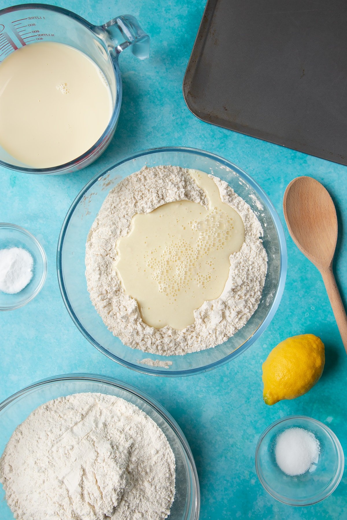 A bowl containing plain flour, salt and bicarbonate of soda, mixed with a well in the centre filled with plant milk. Ingredients to make vegan soda bread surround the bowl.