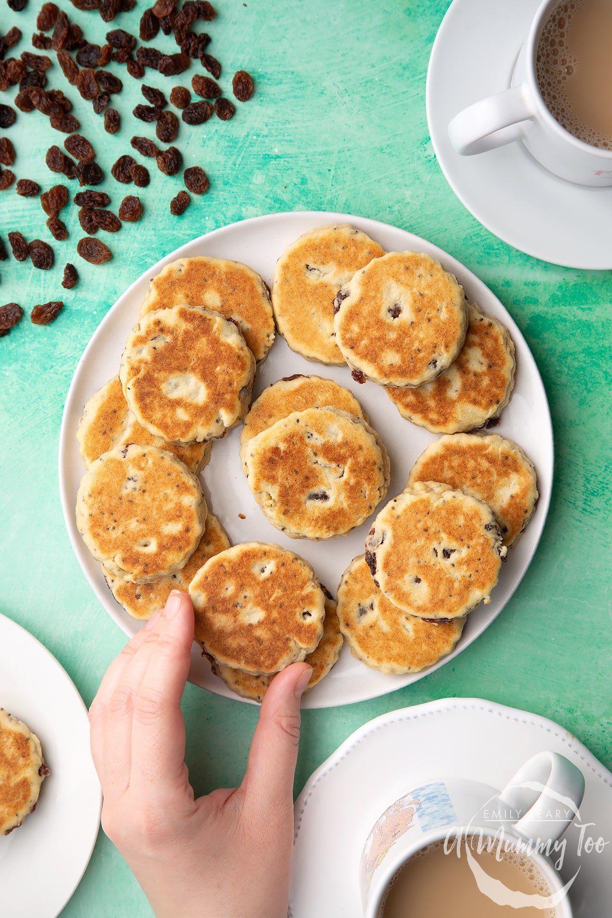 Vegan Welsh cakes arranged on a white plate. Surrounding the plate are cups of tea. A hand reaches in to take a Welsh cake.