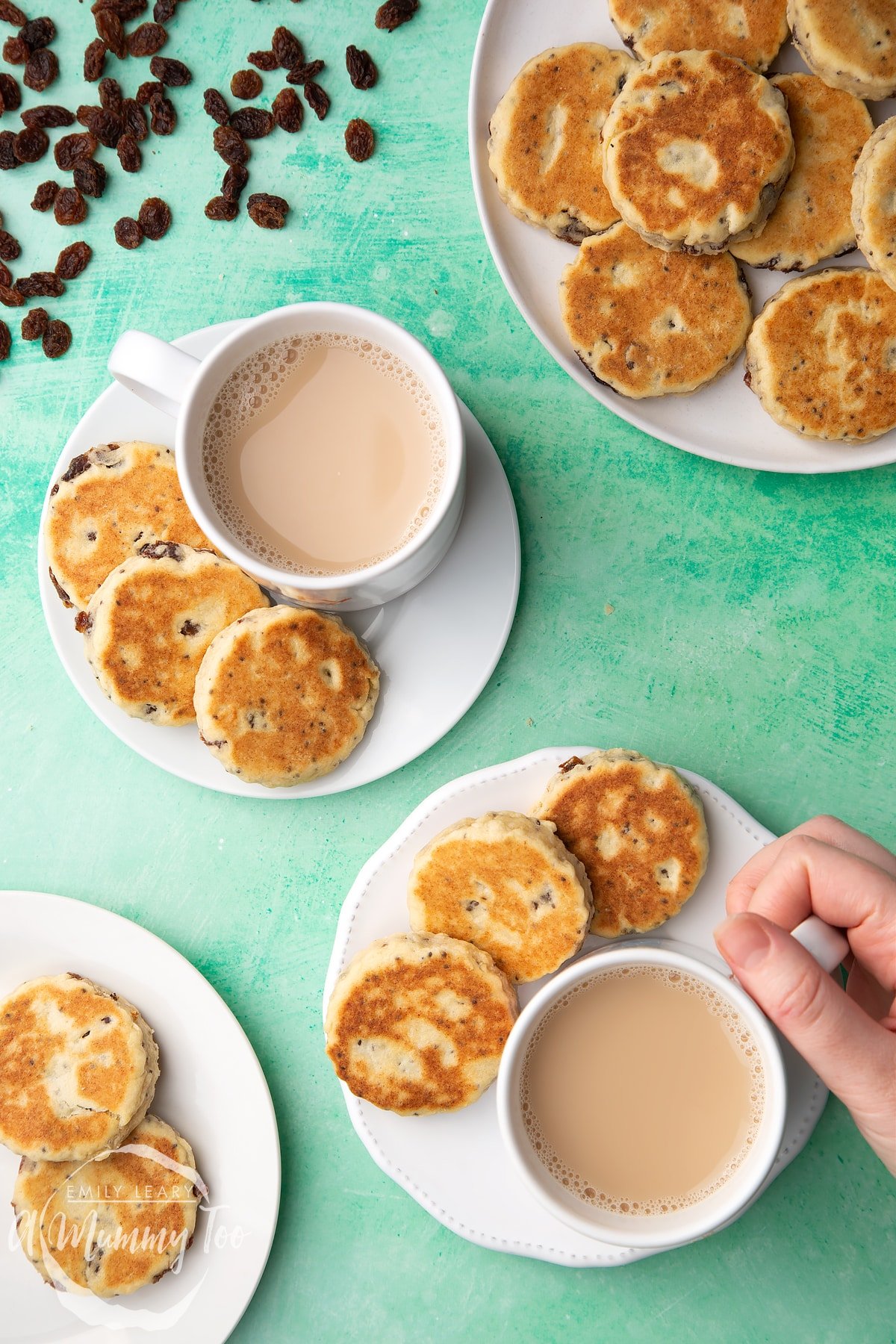 Vegan Welsh cakes arranged on a white plate with cups of tea. A hand reaches in to take a cup.