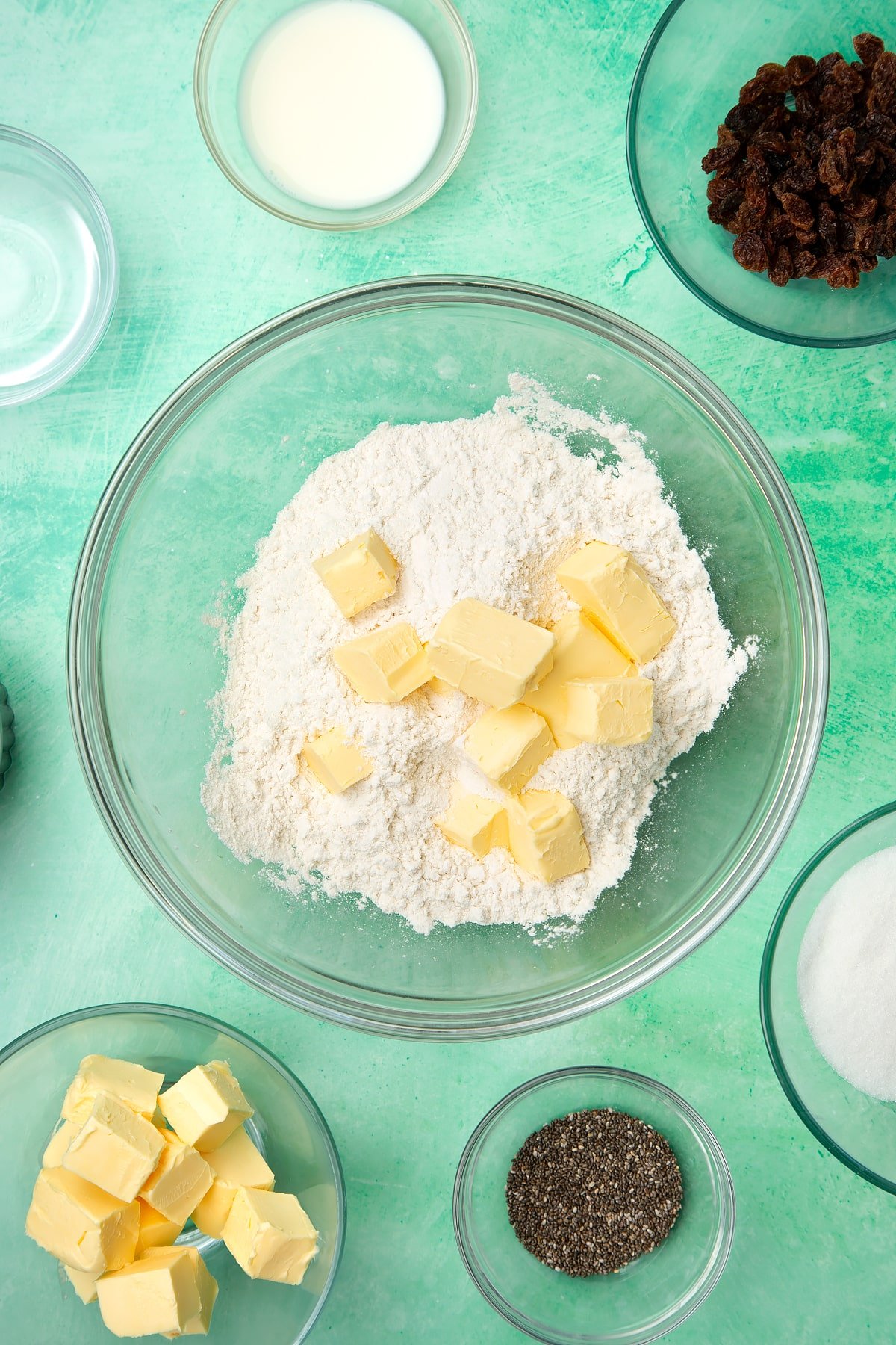 A glass mixing bowl containing self-raising flour and vegan margarine. Ingredients to make vegan Welsh cakes surround the bowl.