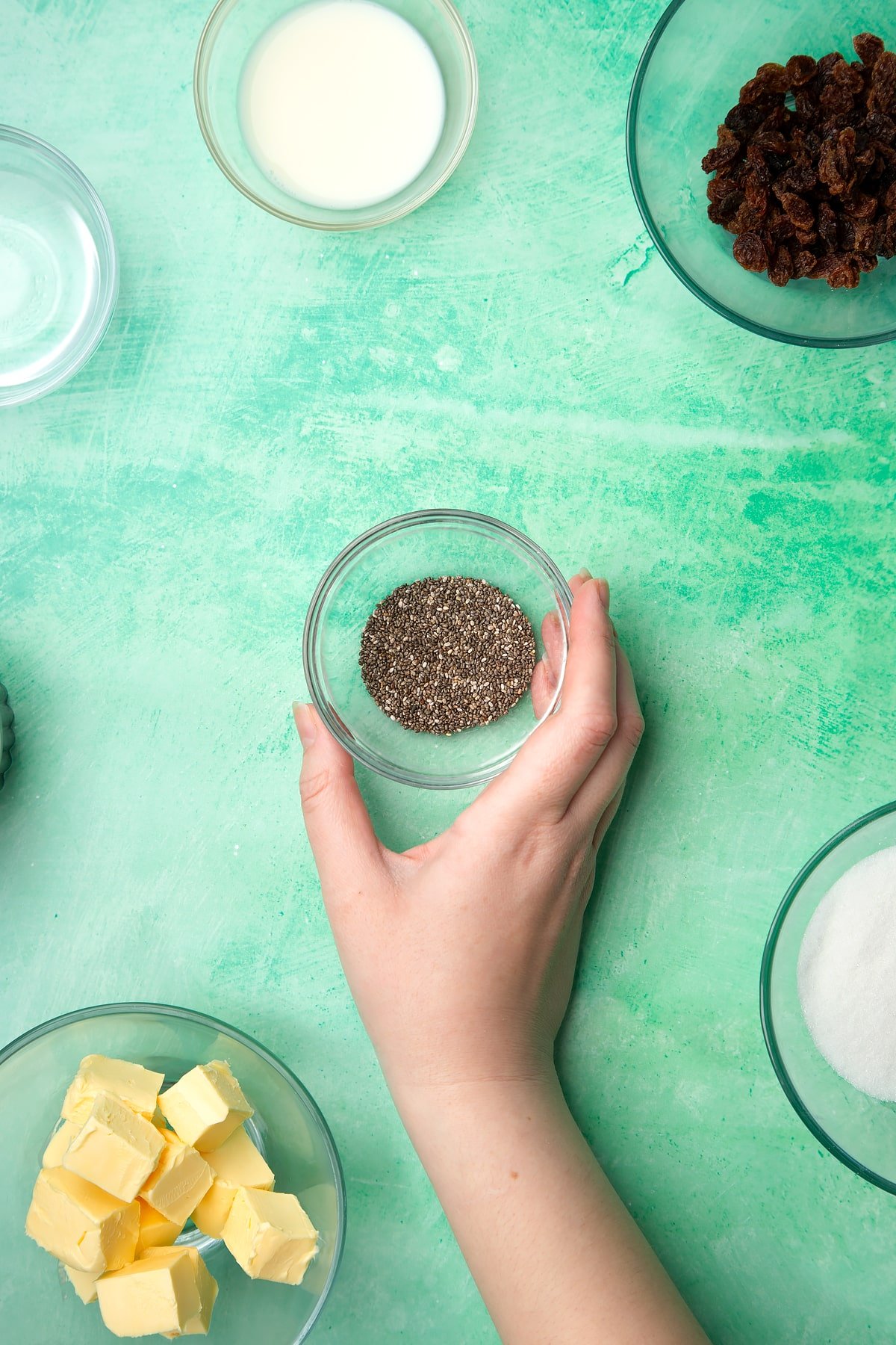 A hand holds a small bowl containing chia seeds. Ingredients to make vegan Welsh cakes surround the bowl.