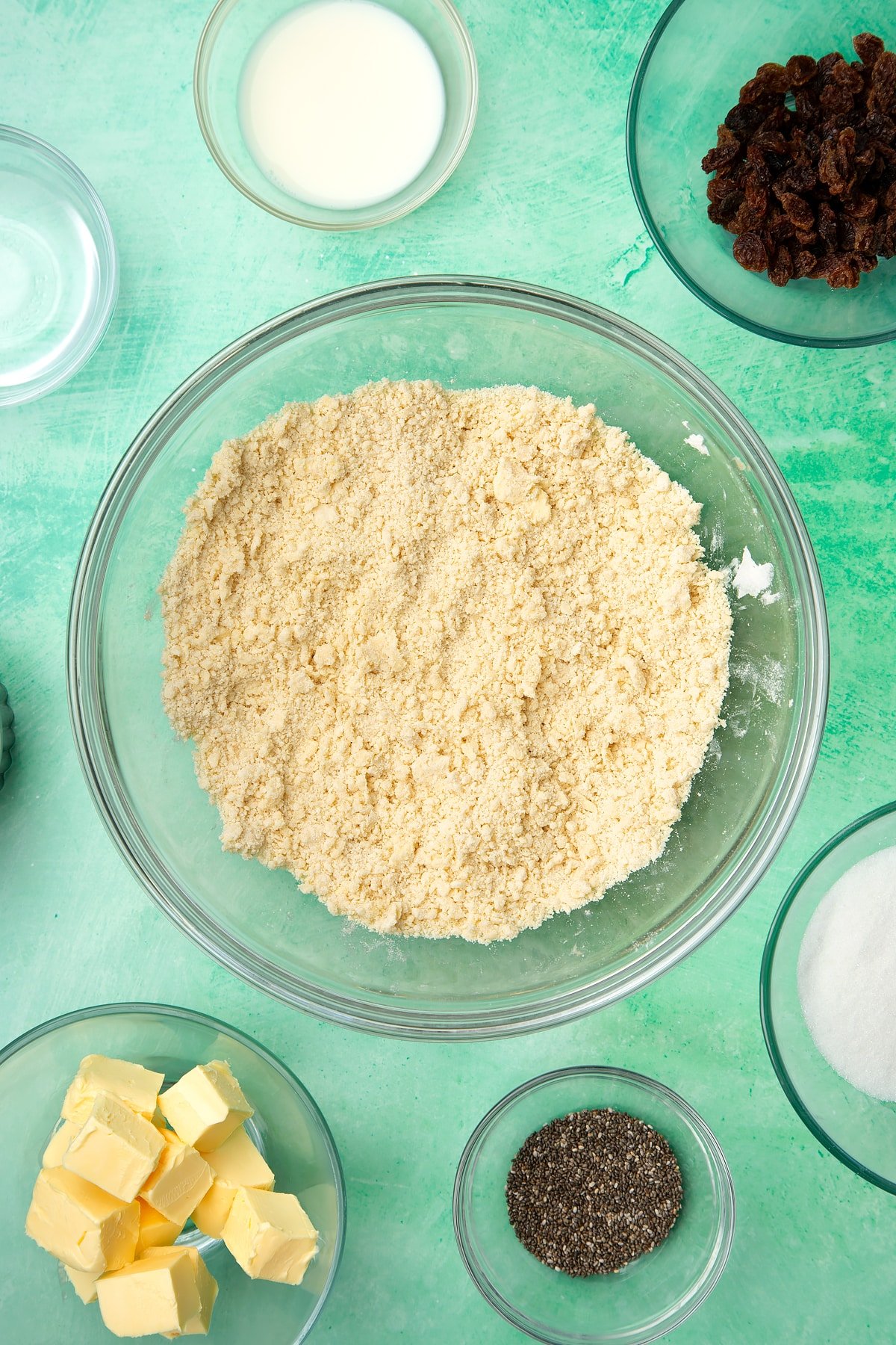 A glass mixing bowl containing self-raising flour and vegan margarine rubbed together. Ingredients to make vegan Welsh cakes surround the bowl.