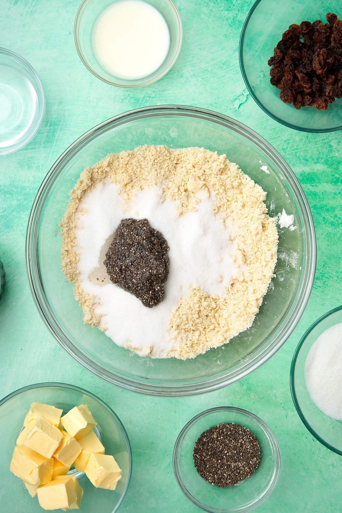 A glass mixing bowl containing self-raising flour and vegan margarine rubbed together, topped with sugar and chia seeds on top. Ingredients to make vegan Welsh cakes surround the bowl.