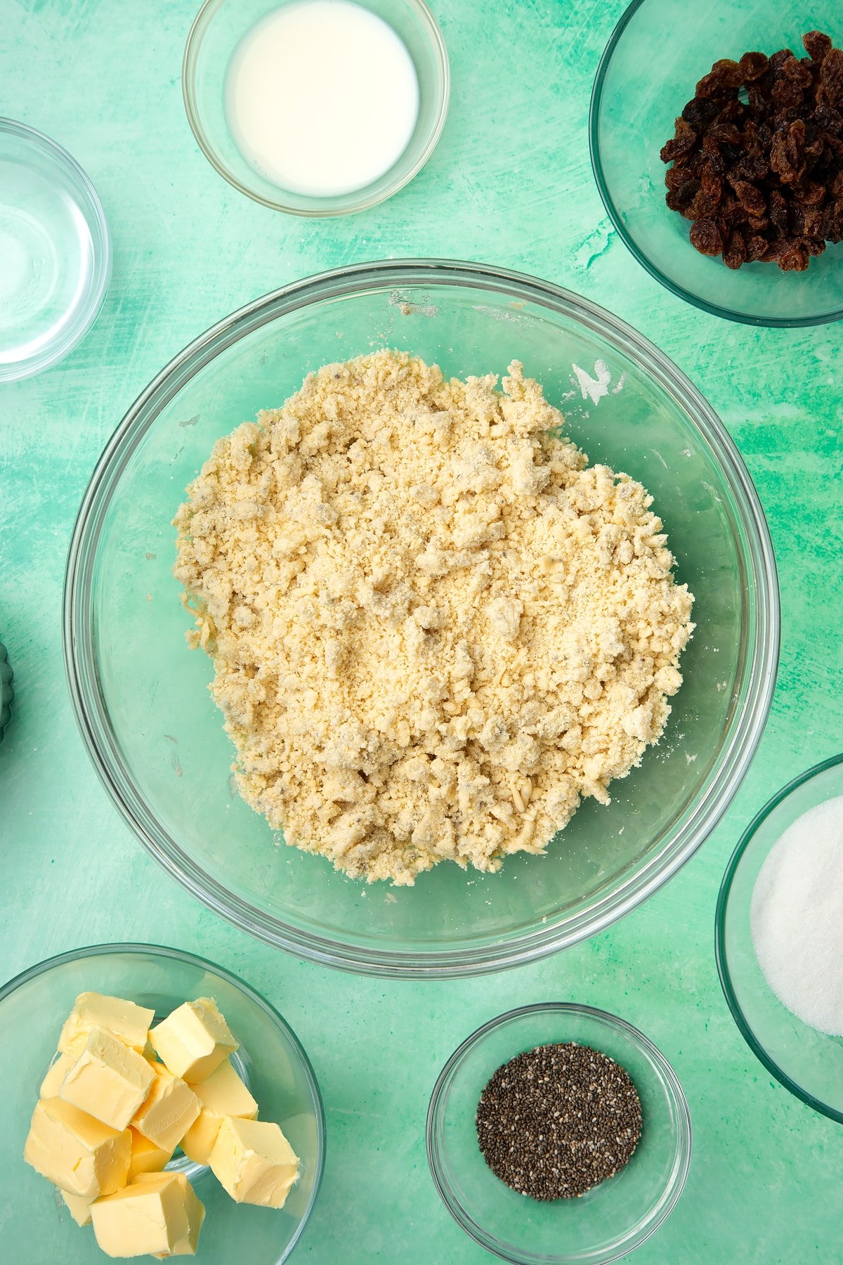 A glass mixing bowl containing self-raising flour, vegan margarine, chia seeds and sugar mixed together. Ingredients to make vegan Welsh cakes surround the bowl.