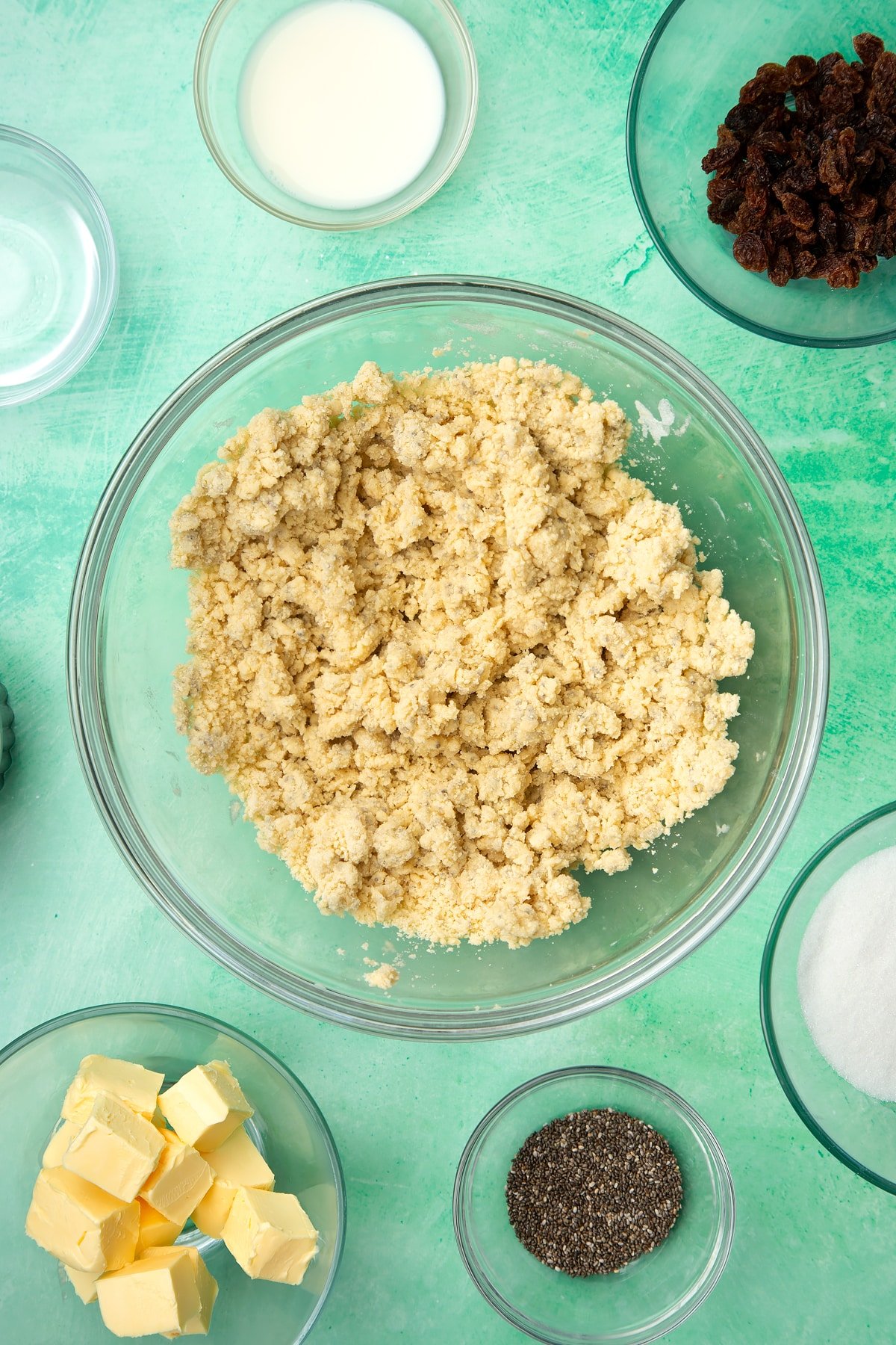 A glass mixing bowl containing self-raising flour, vegan margarine, chia seeds, sugar and plant milk mixed together. Ingredients to make vegan Welsh cakes surround the bowl.