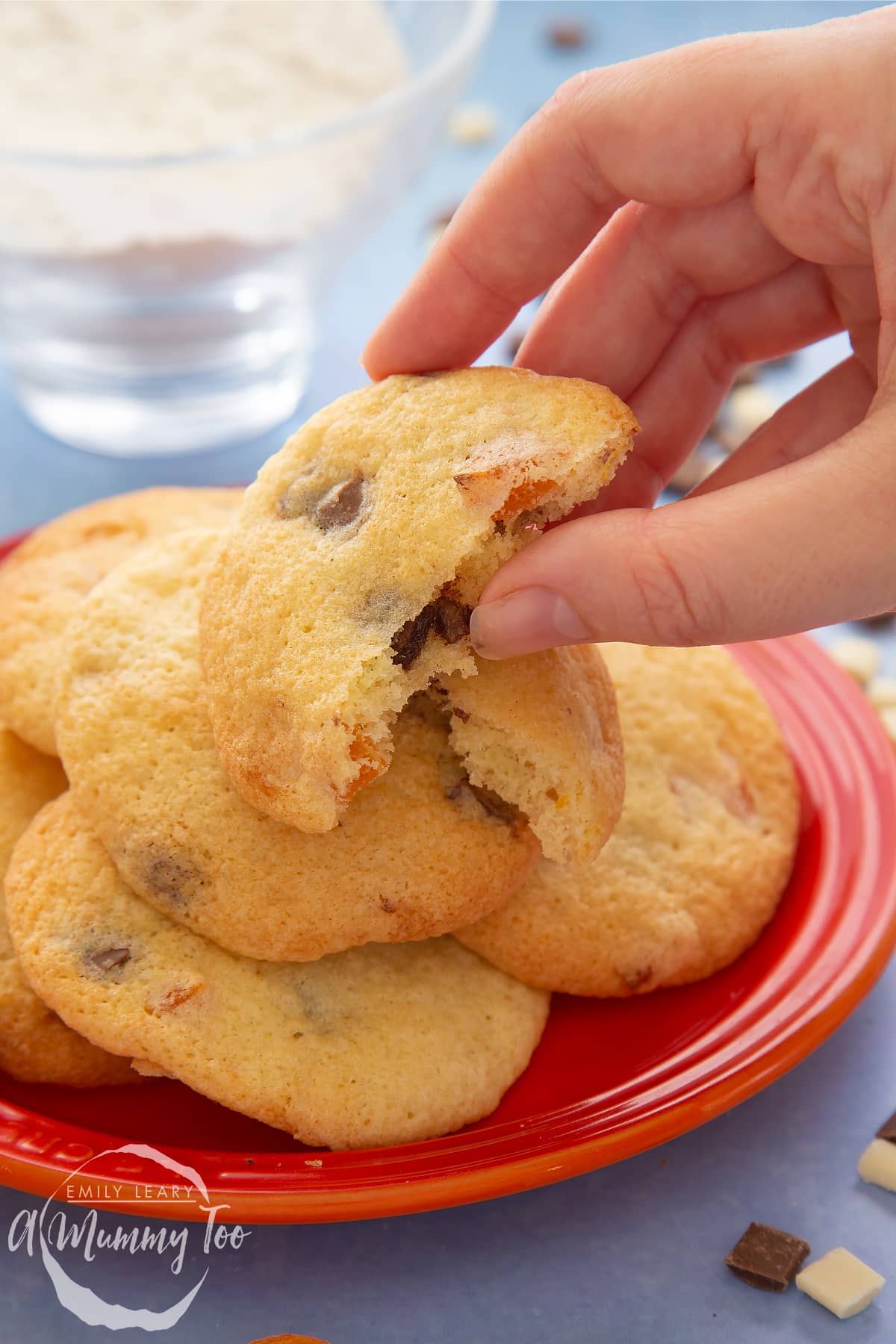 Apricot chocolate chip cookies stacked on an orange plate. The top cookie is broken in two. A hand reaches to take a piece.