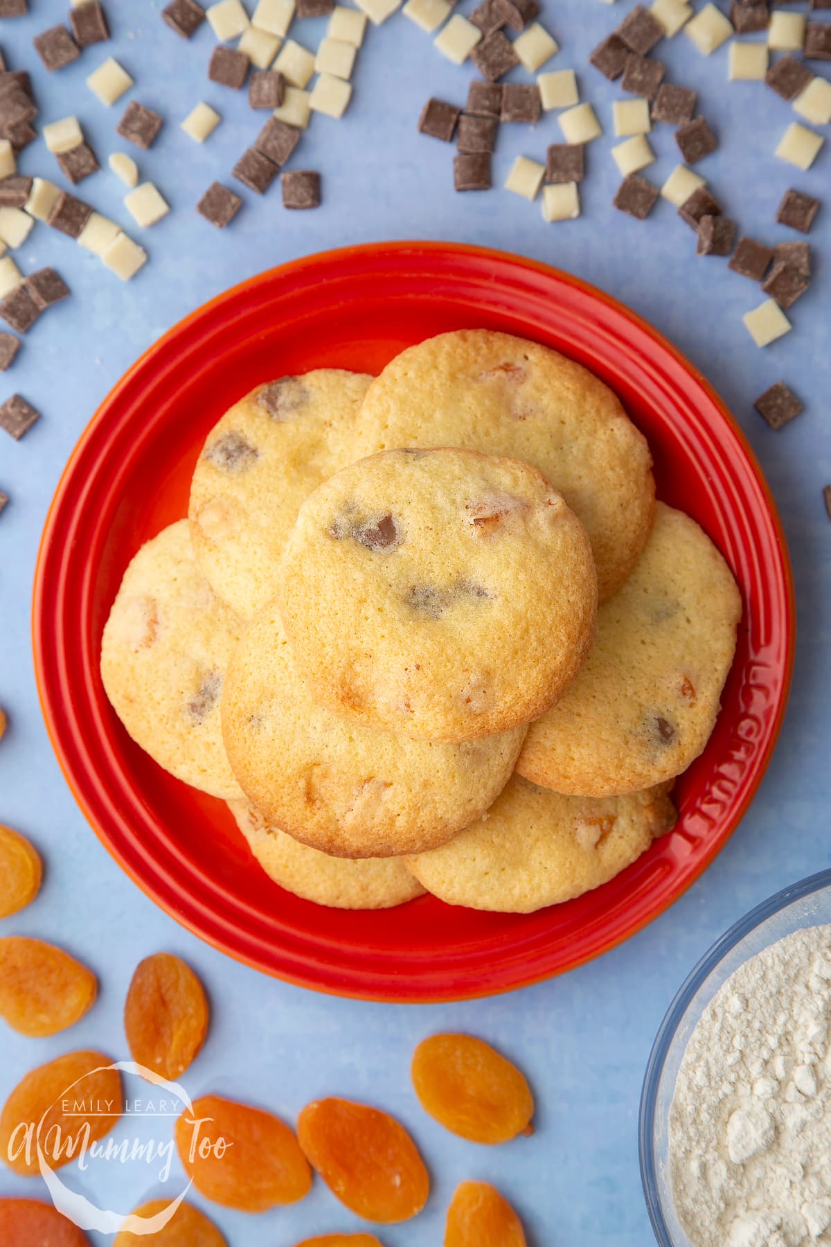 Apricot chocolate chip cookies stacked on a small orange plate. Chocolate chips and dried apricots are scattered around.