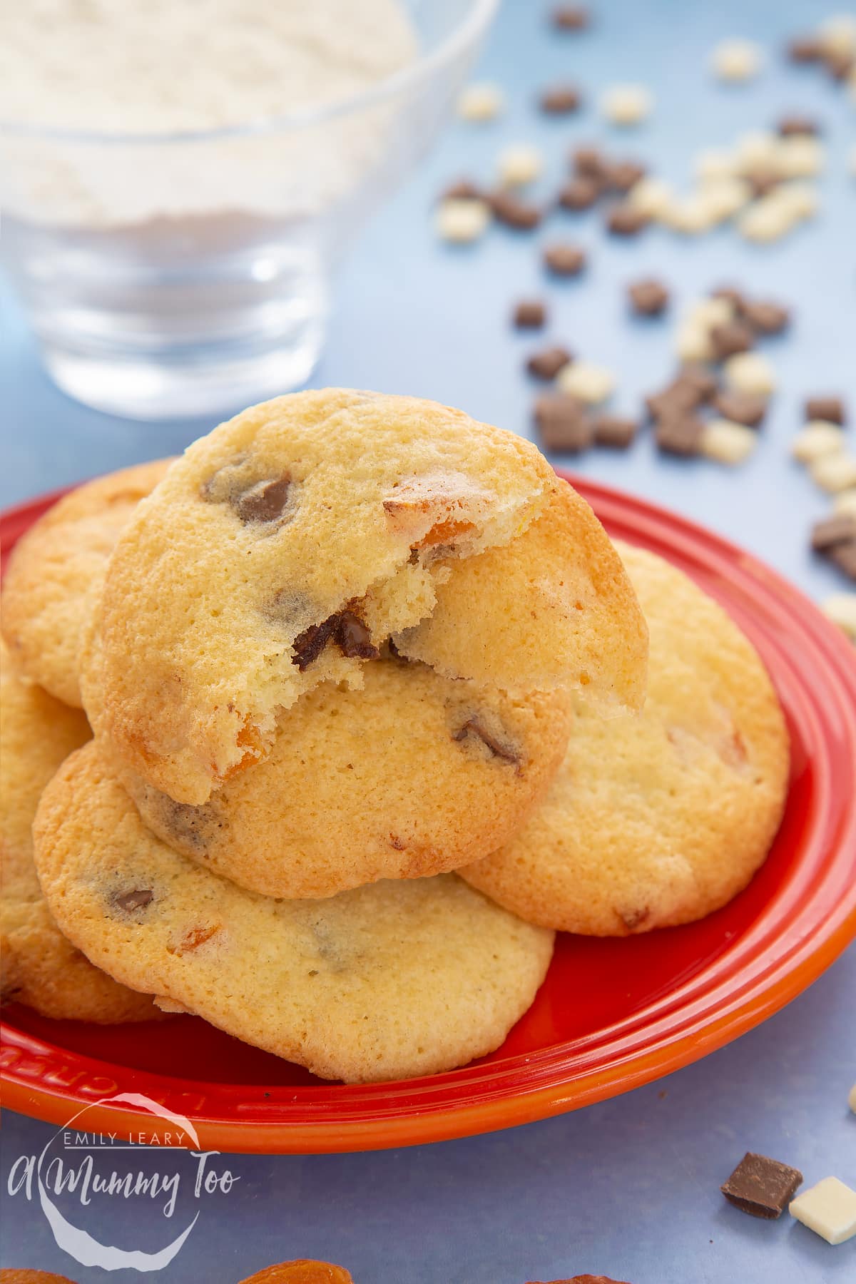 Apricot chocolate chip cookies stacked on a small orange plate. Chocolate chips and dried apricots are scattered around. One cookie is broken in two.