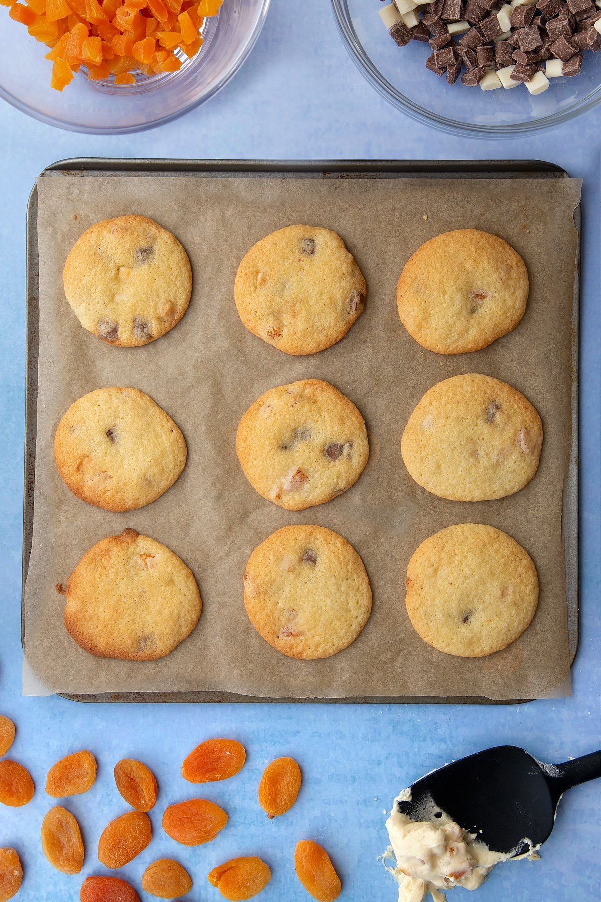 Freshly baked chocolate and apricot cookies on a tray lined with baking paper. Ingredients to make apricot chocolate chip cookies surround the tray.