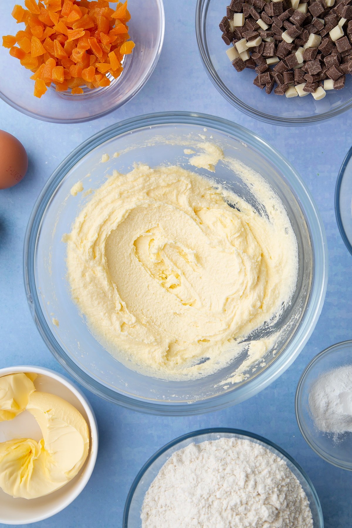 Margarine and sugar creamed together in a glass mixing bowl. Ingredients to make apricot chocolate chip cookies surround the bowl.