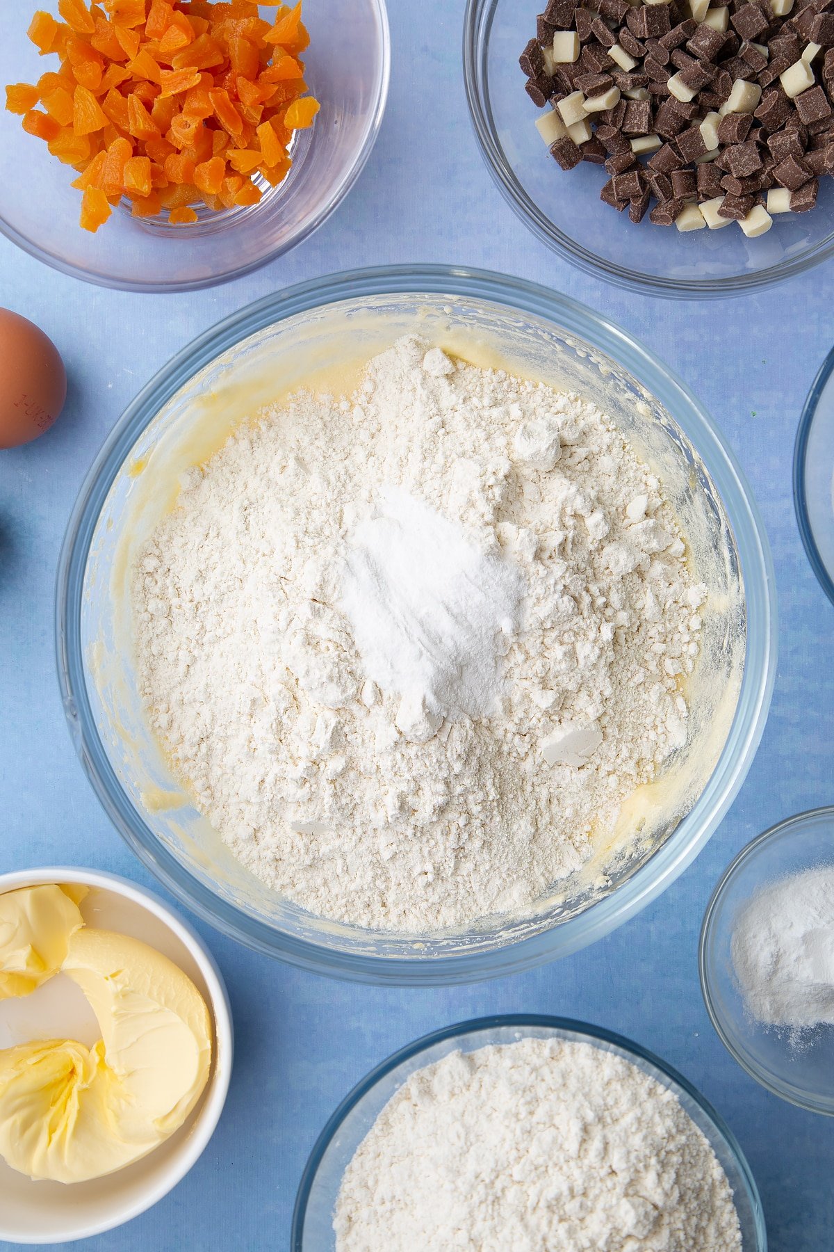 Margarine, sugar and eggs creamed together in a glass mixing bowl with flour and baking powder on top. Ingredients to make apricot chocolate chip cookies surround the bowl.