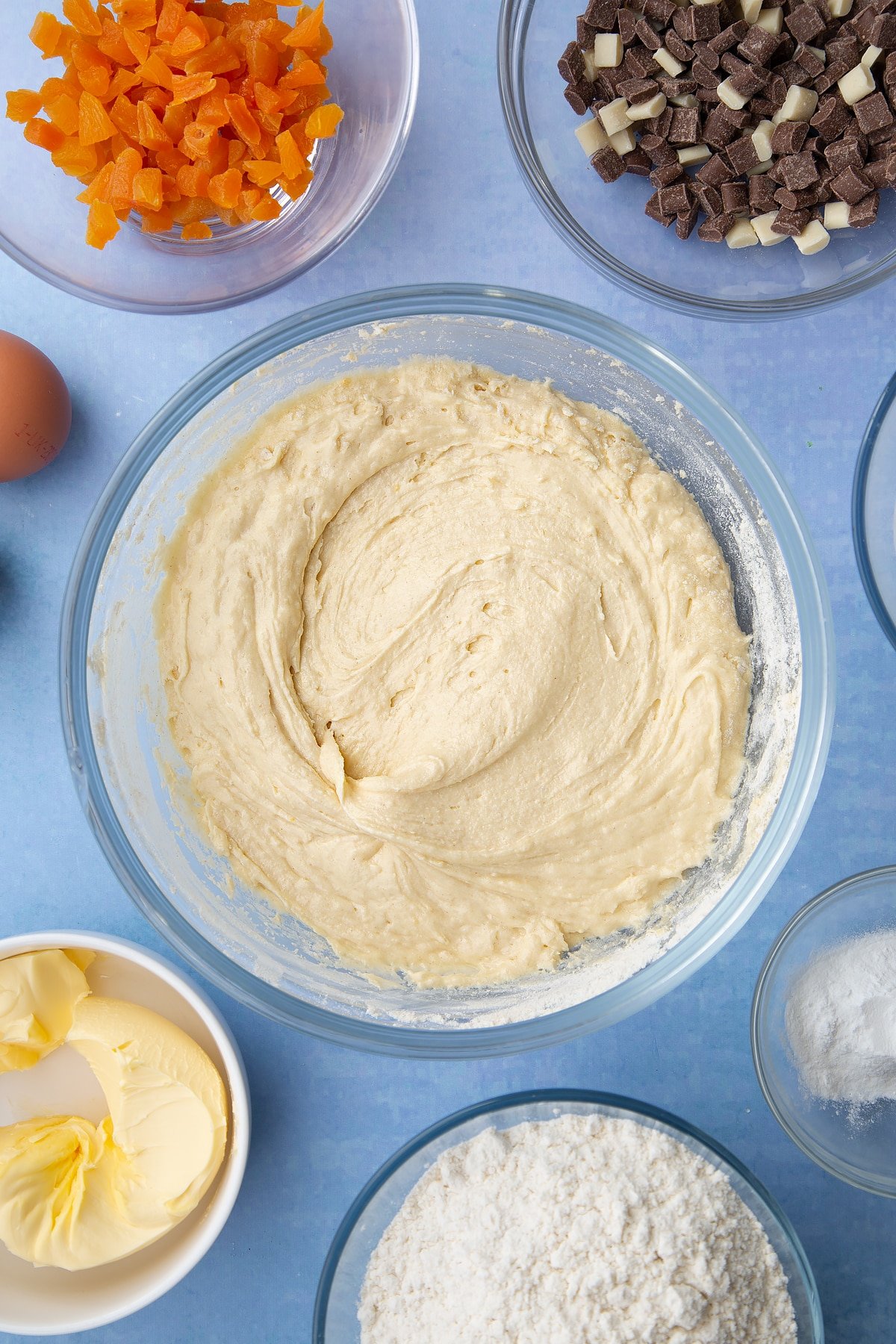 Cookie dough in a glass mixing bowl. Ingredients to make apricot chocolate chip cookies surround the bowl.