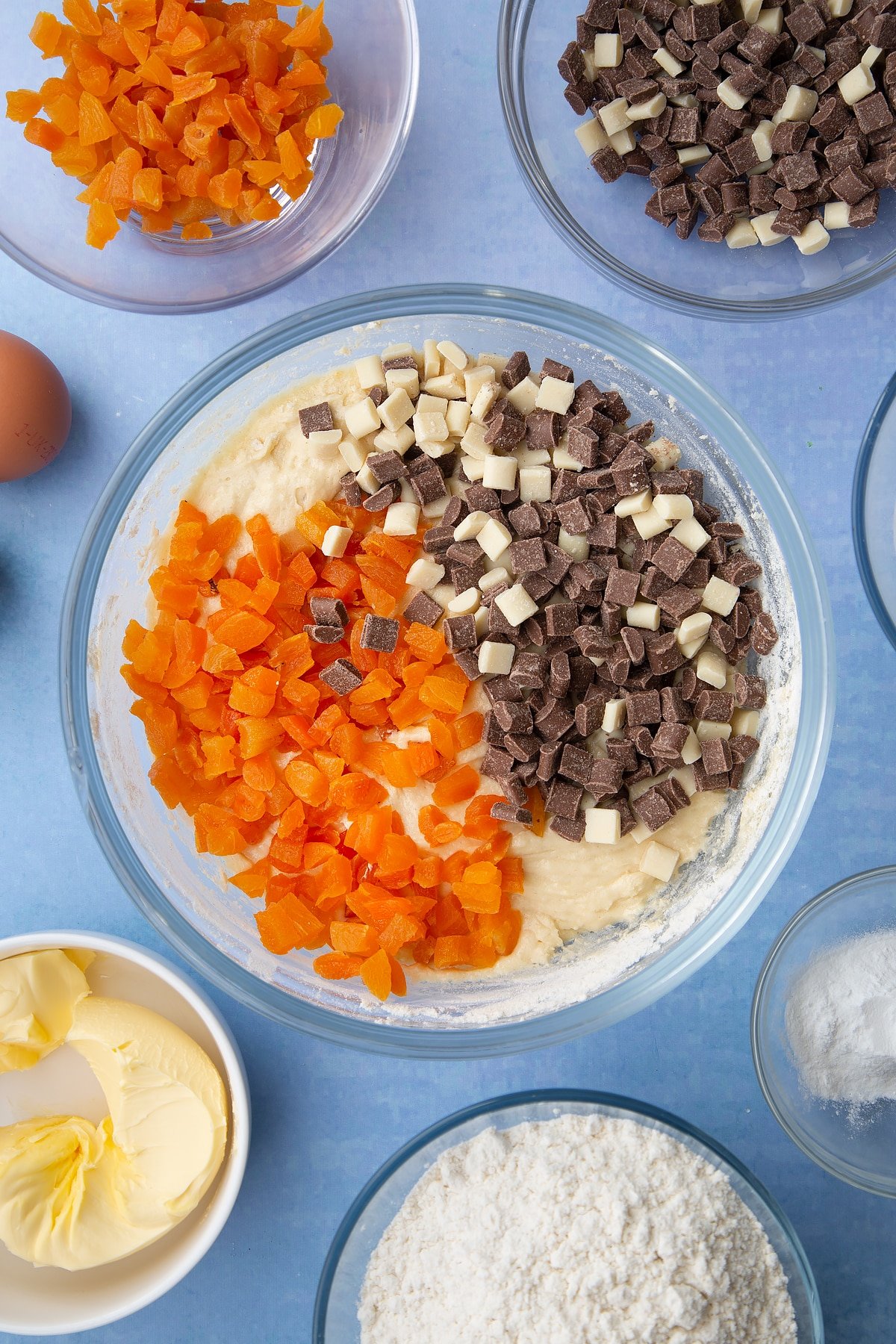 Cookie dough in a glass mixing bowl with chopped dried apricots and chocolate chips on top. Ingredients to make apricot chocolate chip cookies surround the bowl.