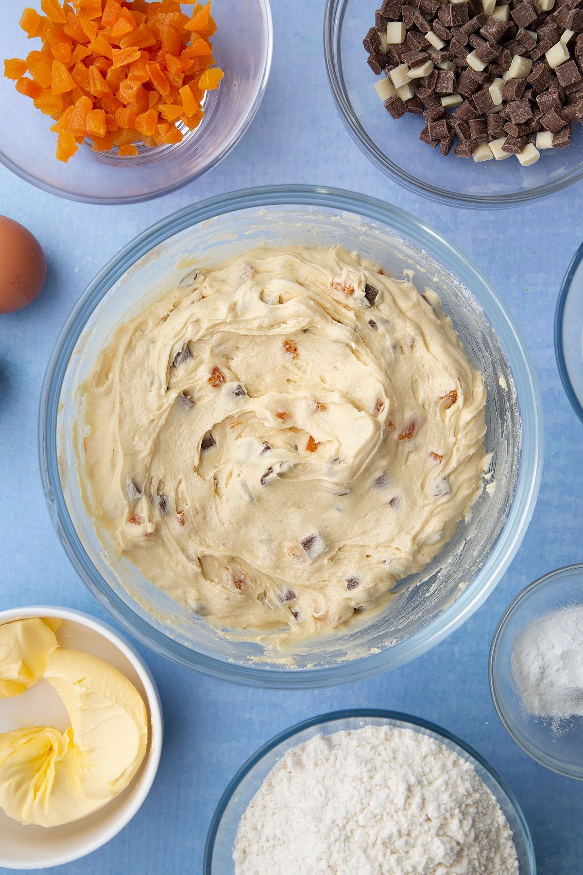 Chocolate and apricot cookie dough in a glass mixing bowl. Ingredients to make apricot chocolate chip cookies surround the bowl.