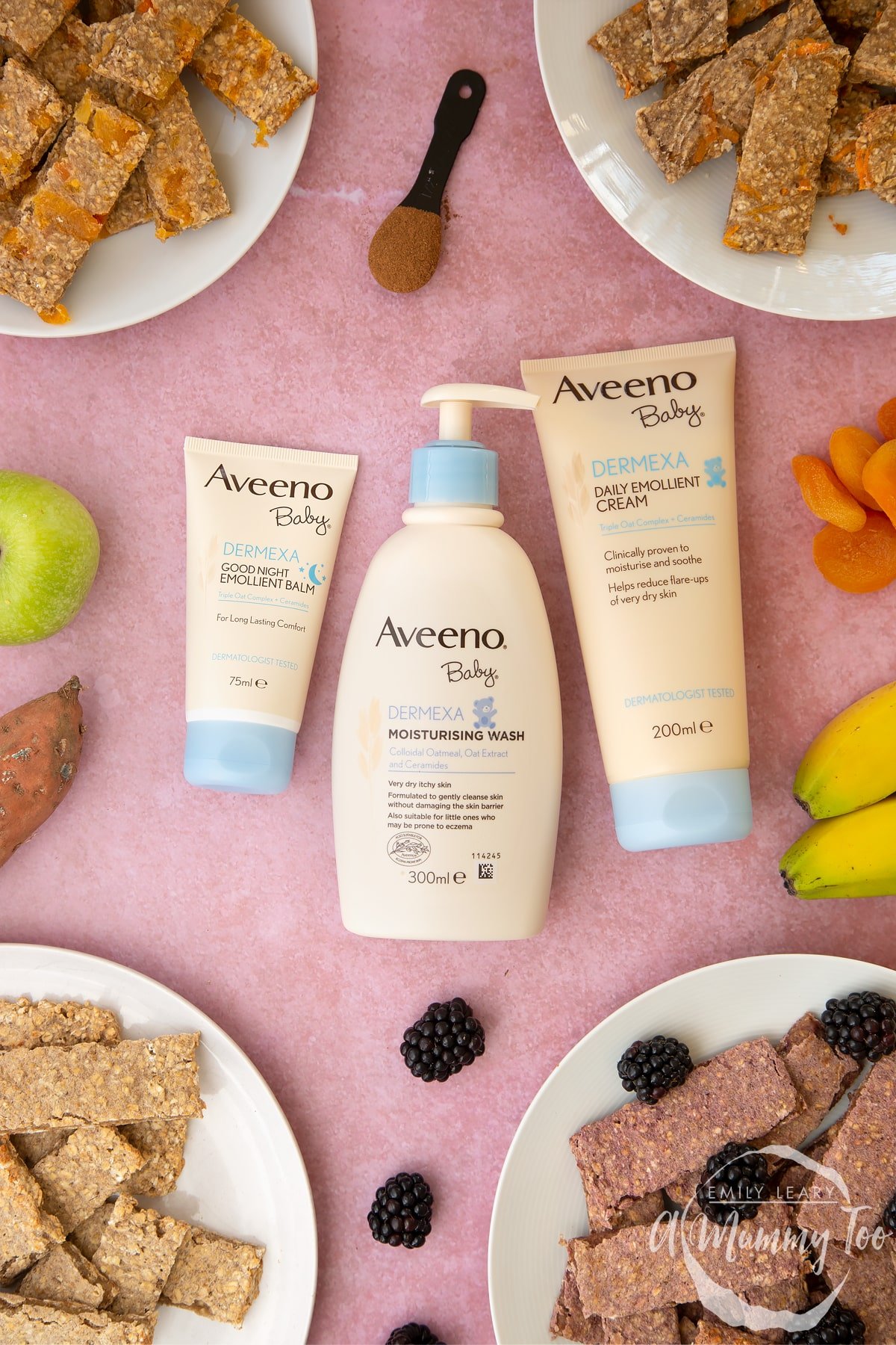 AVEENO Baby skincare products laying on a pink surface. Various flavours of oat fingers sit on plates around the edge of the frame.