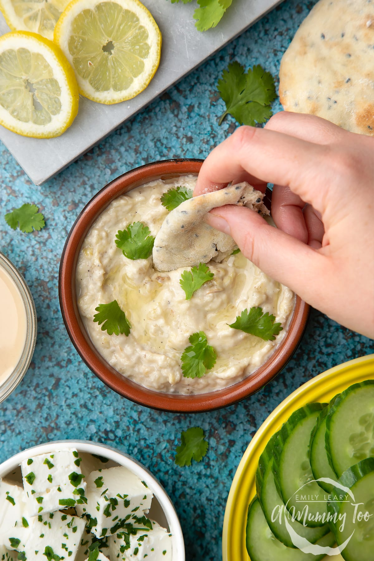 Baba ganoush in a shallow terracotta dish. Feta, cucumbers, lemon and flat bread surround the dish. A hand dips flat bread into the baba ganoush.