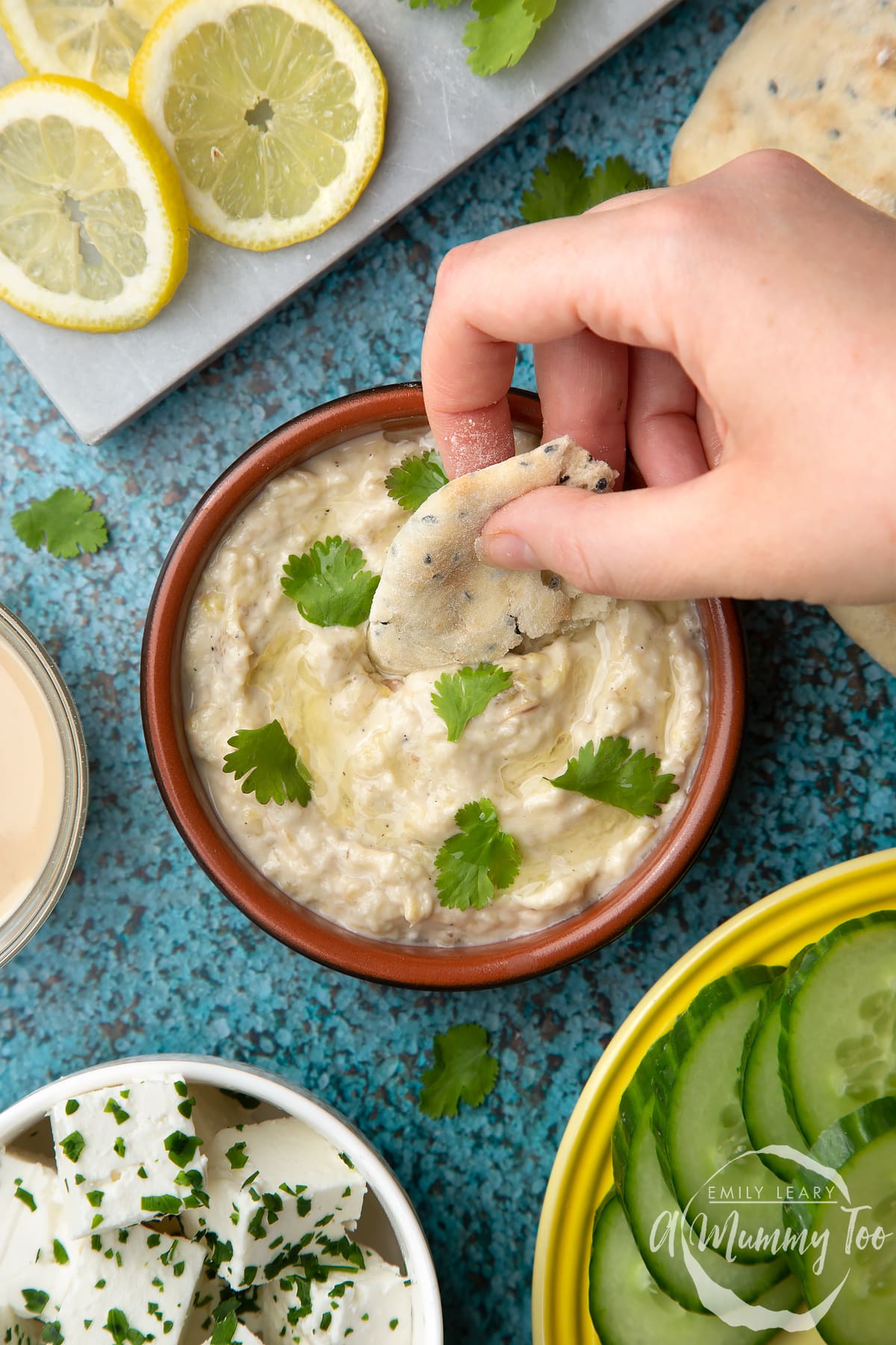 Baba ganoush in a shallow terracotta dish. A hand dips flat bread into the dish. Feta, cucumbers, lemon and flat bread surround the dish.