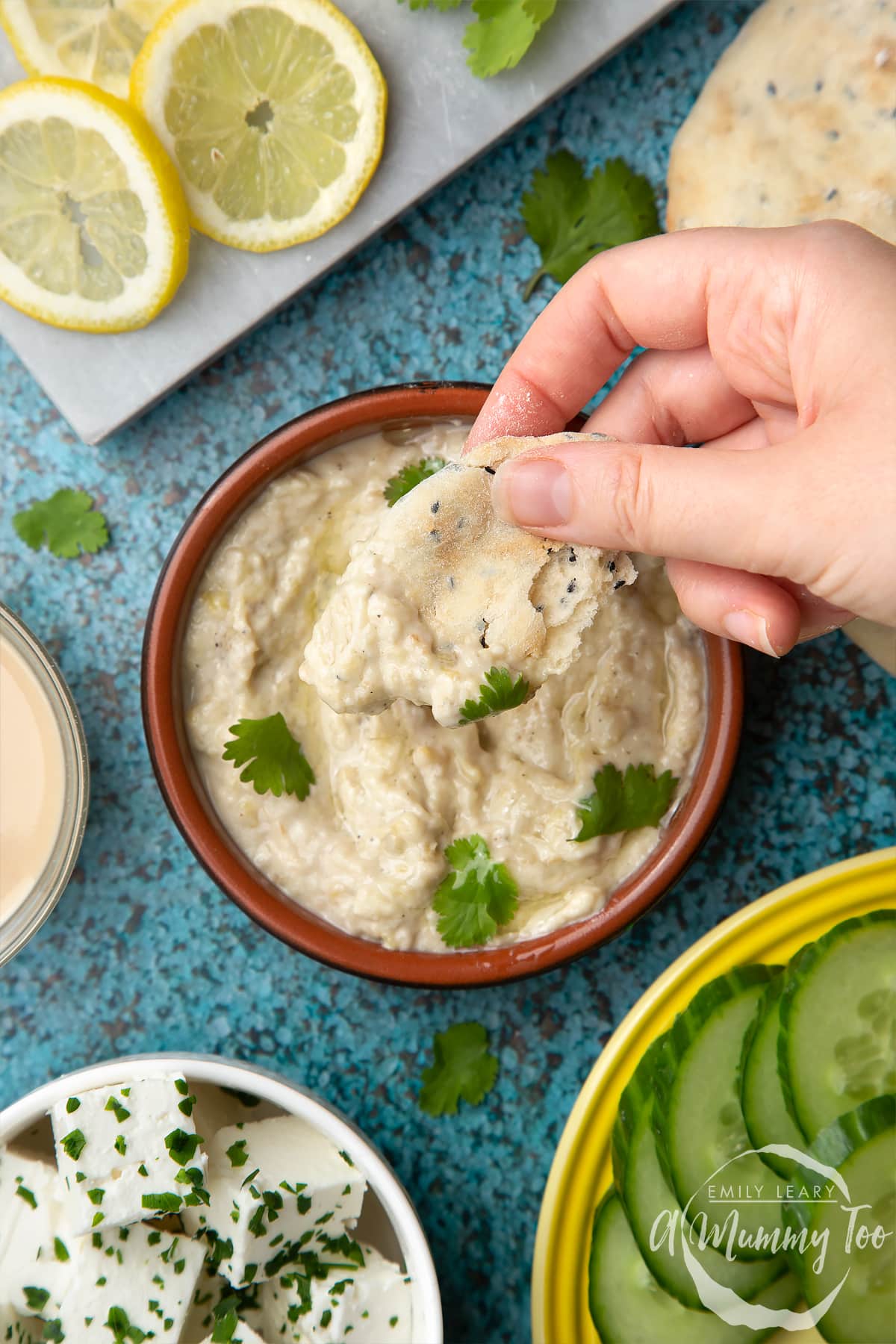 Baba ganoush in a small, shallow teracotta dish. The dip has been garnished with olive oil and parsley leaves. A hand holds a piece of flatbread above the bowl.