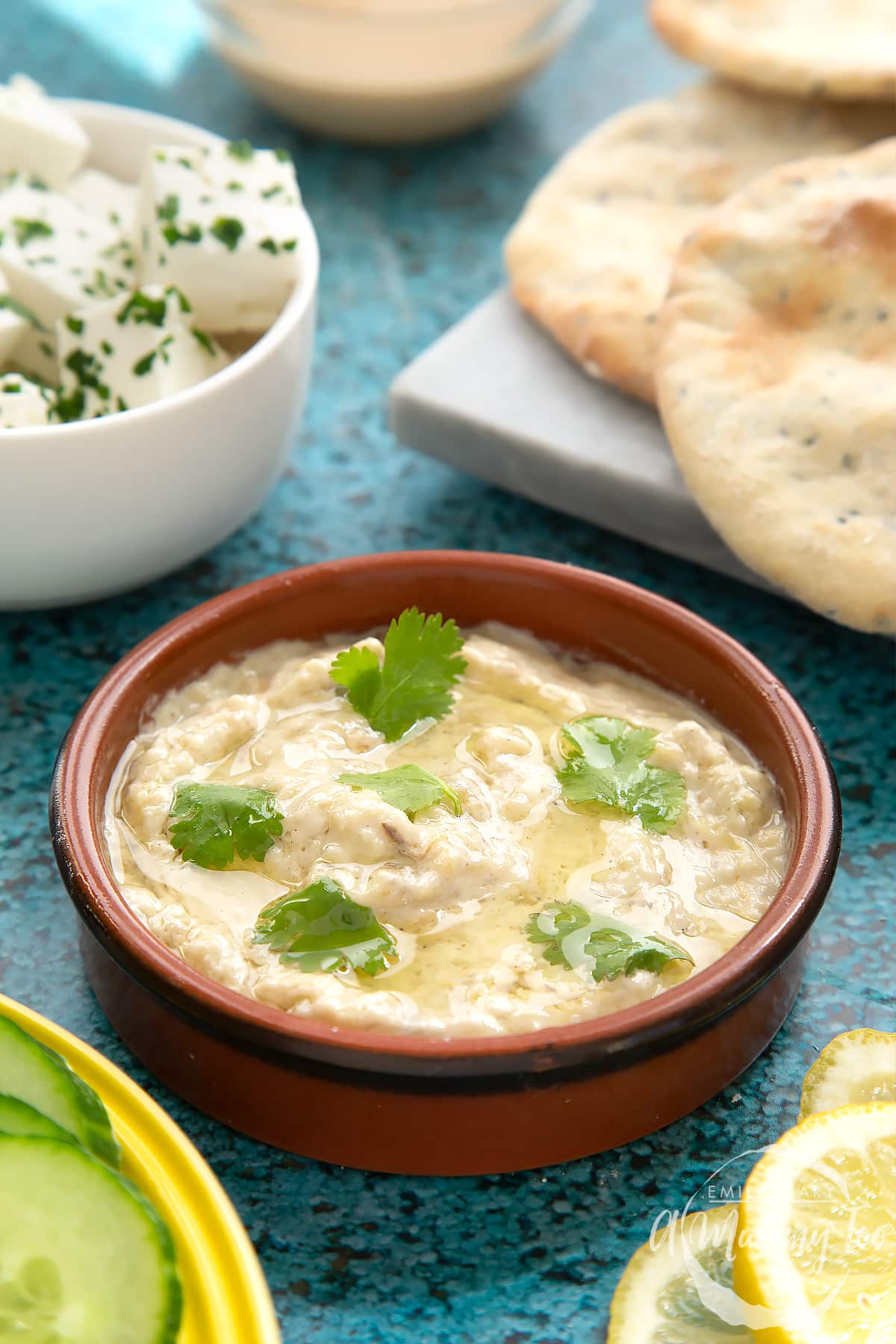 Baba ganoush in a shallow terracotta dish. Feta, cucumbers, lemon and flat bread surround the dish.