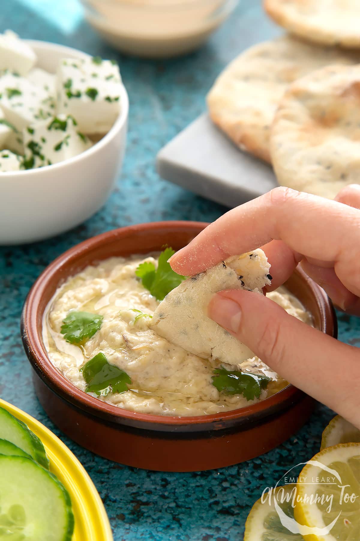 Baba ganoush in a small, shallow teracotta dish. The dip has been garnished with olive oil and parsley leaves. A hand dips in some flatbread.