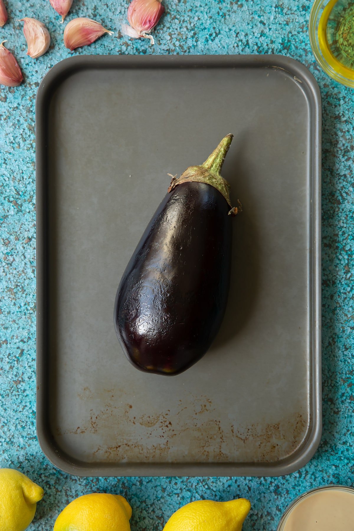 An aubergine on a baking tray. Ingredients to make an easy baba ganoush recipe surround the tray. 