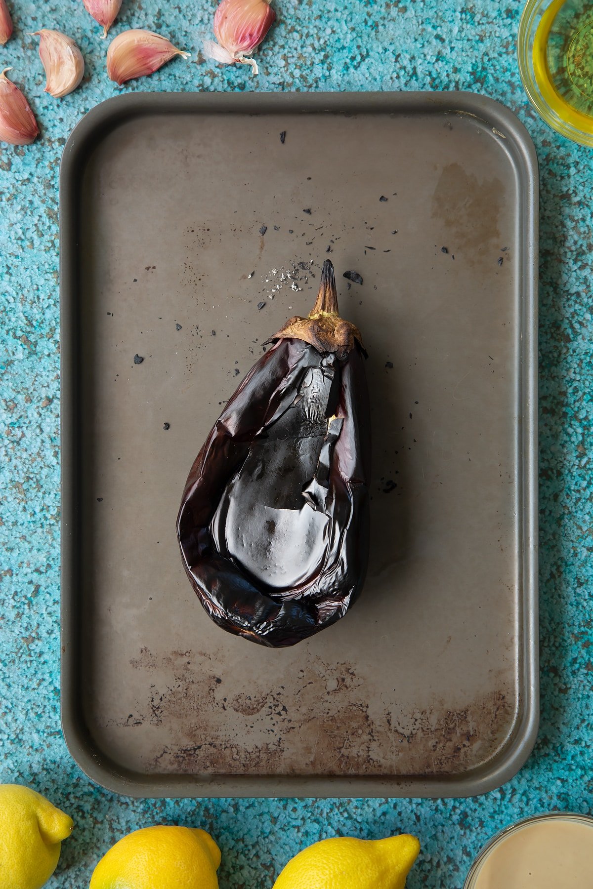 A charred aubergine on a baking tray. Ingredients to make an easy baba ganoush recipe surround the tray. 