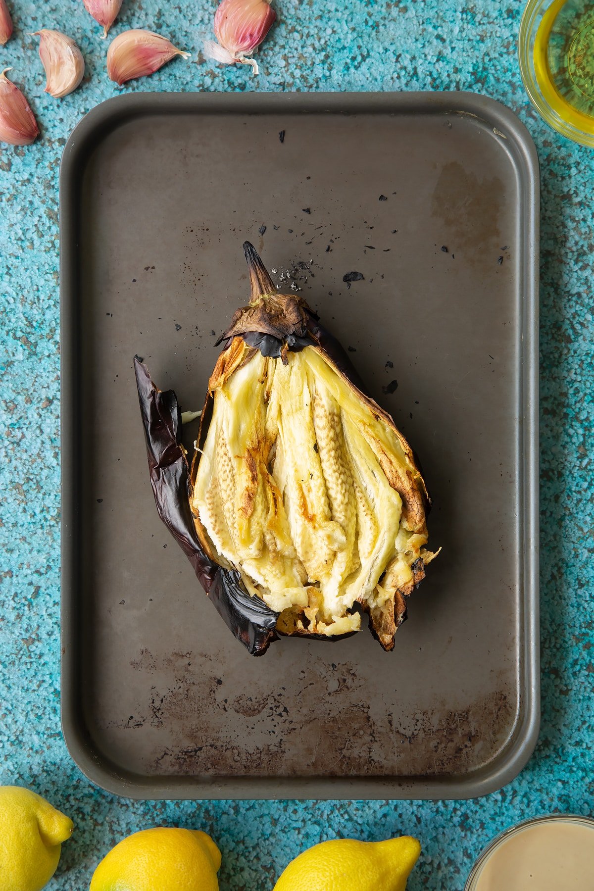 A charred aubergine on a baking tray. It has been torn open to reveal the flesh inside. Ingredients to make an easy baba ganoush recipe surround the tray. 