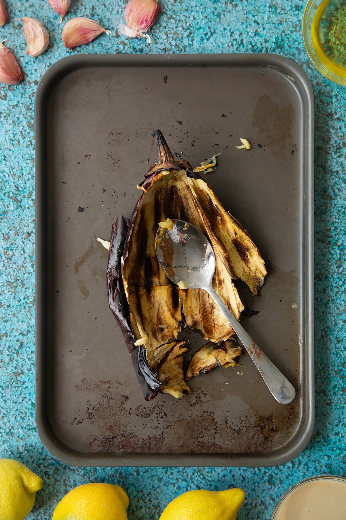 A charred aubergine skin on a baking tray with a spoon. Ingredients to make an easy baba ganoush recipe surround the tray. 