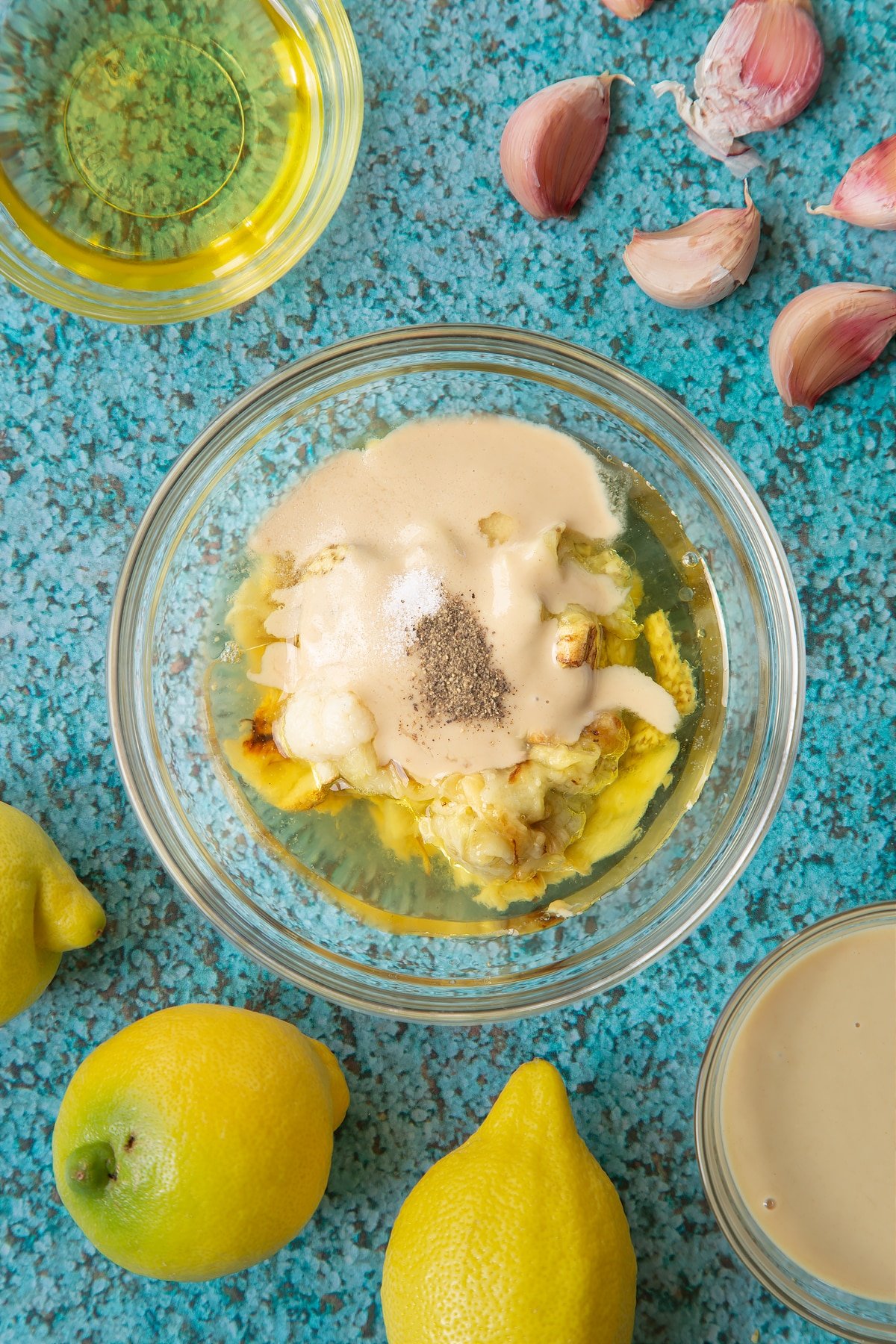 Aubergine flesh in a small glass bowl with tahini paste, olive oil, lemon juice, minced garlic, salt and pepper. Ingredients to make an easy baba ganoush recipe surround the bowl. 