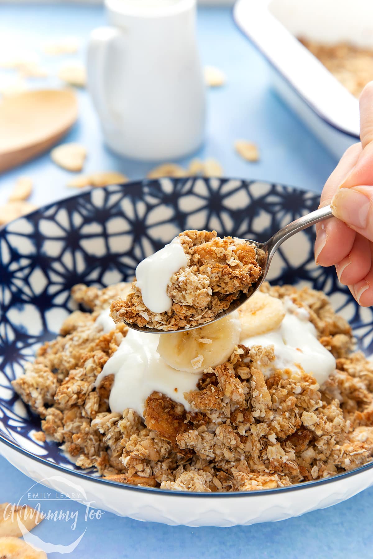 Banana coconut granola in a bowl, topped with yogurt and fresh banana slices. A hand holds a spoon filled with granola.