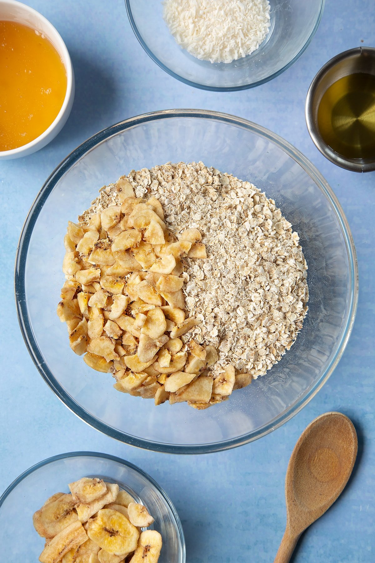  A large glass mixing bowl containing rolled oats and dried banana chips. Ingredients to make a banana coconut granola recipe surround the bowl.