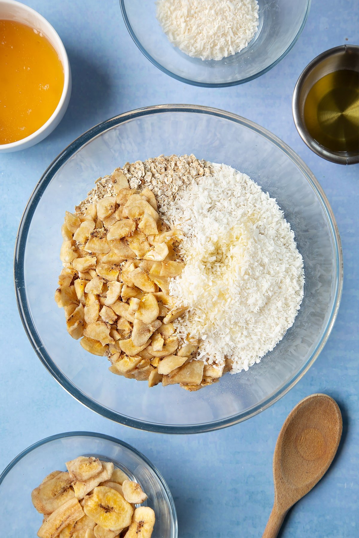  A large glass mixing bowl containing rolled oats, dried banana chips, desiccated coconut and oil. Ingredients to make a banana coconut granola recipe surround the bowl.