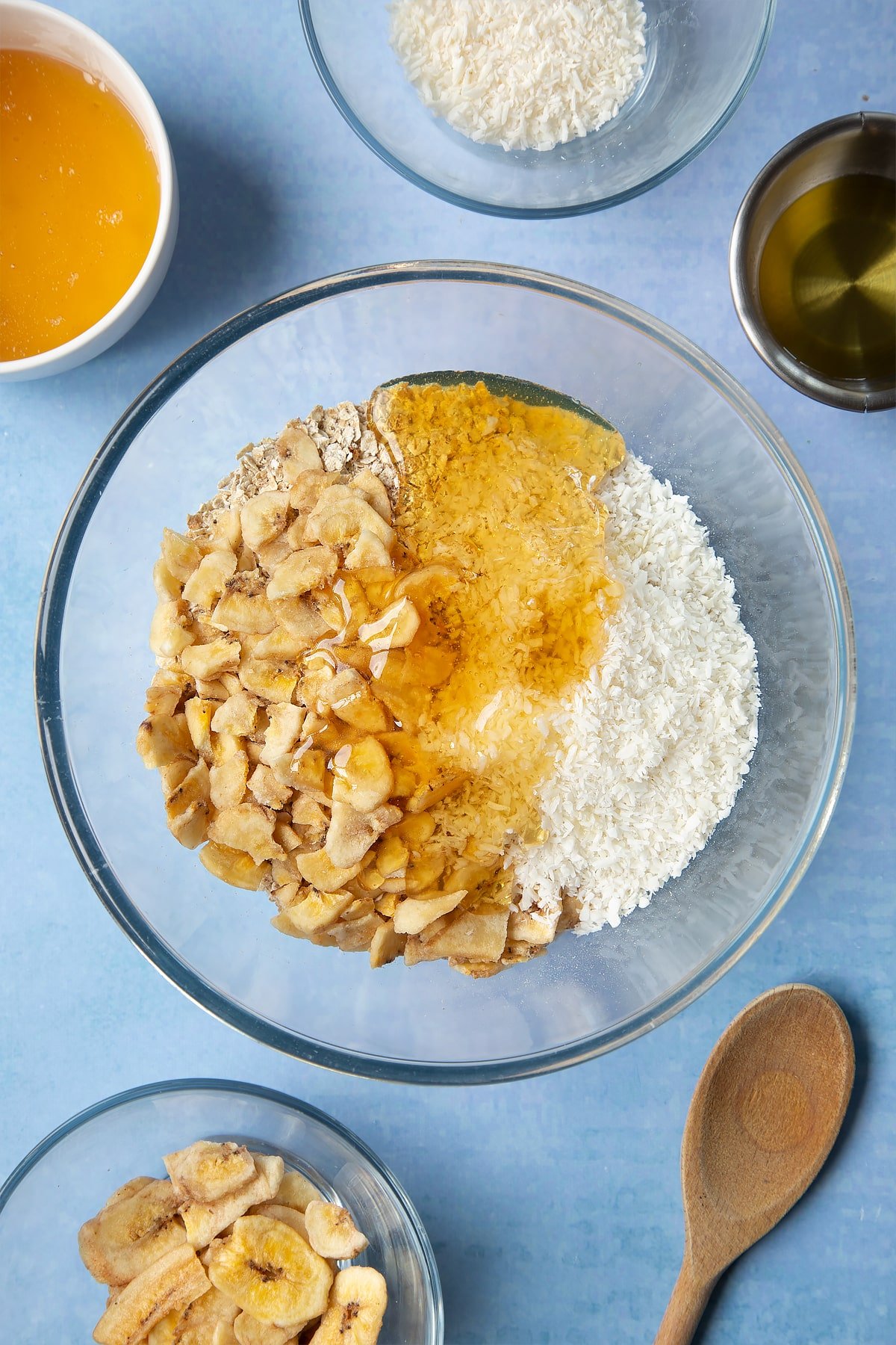  A large glass mixing bowl containing rolled oats, dried banana chips, desiccated coconut, oil and honey. Ingredients to make a banana coconut granola recipe surround the bowl.