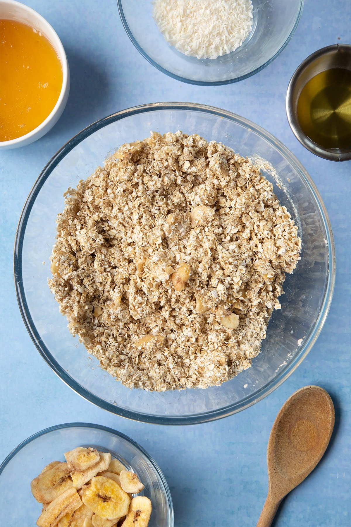  A large glass mixing bowl containing rolled oats, dried banana chips, desiccated coconut, oil and honey mixed together. Ingredients to make a banana coconut granola recipe surround the bowl.