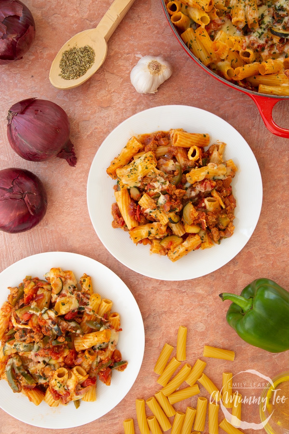 Bolognese al forno served onto two white plates. Ingredients surround the plates. A casserole dish containing the pasta bake is shown to the side.