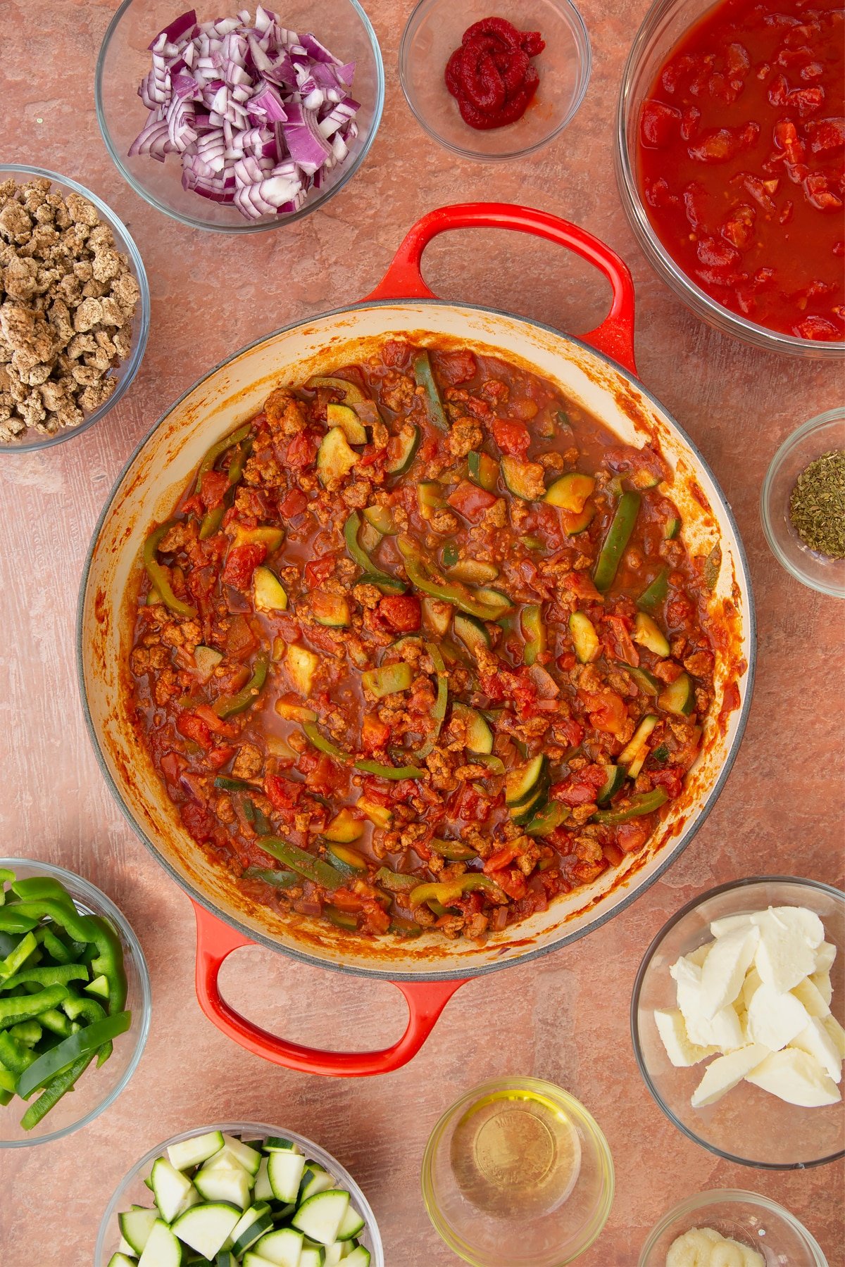 A casserole dish containing a bolognese sauce of red onion, garlic, sliced peppers, courgettes, mince, chopped tomatoes, tomato puree and dried oregano. Ingredients to make bolognese al forno surround the dish.