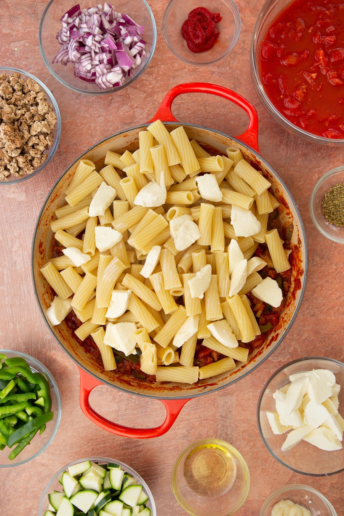 A casserole dish containing a bolognese sauce topped with cooked pasta and torn mozzarella. Ingredients to make bolognese al forno surround the dish.