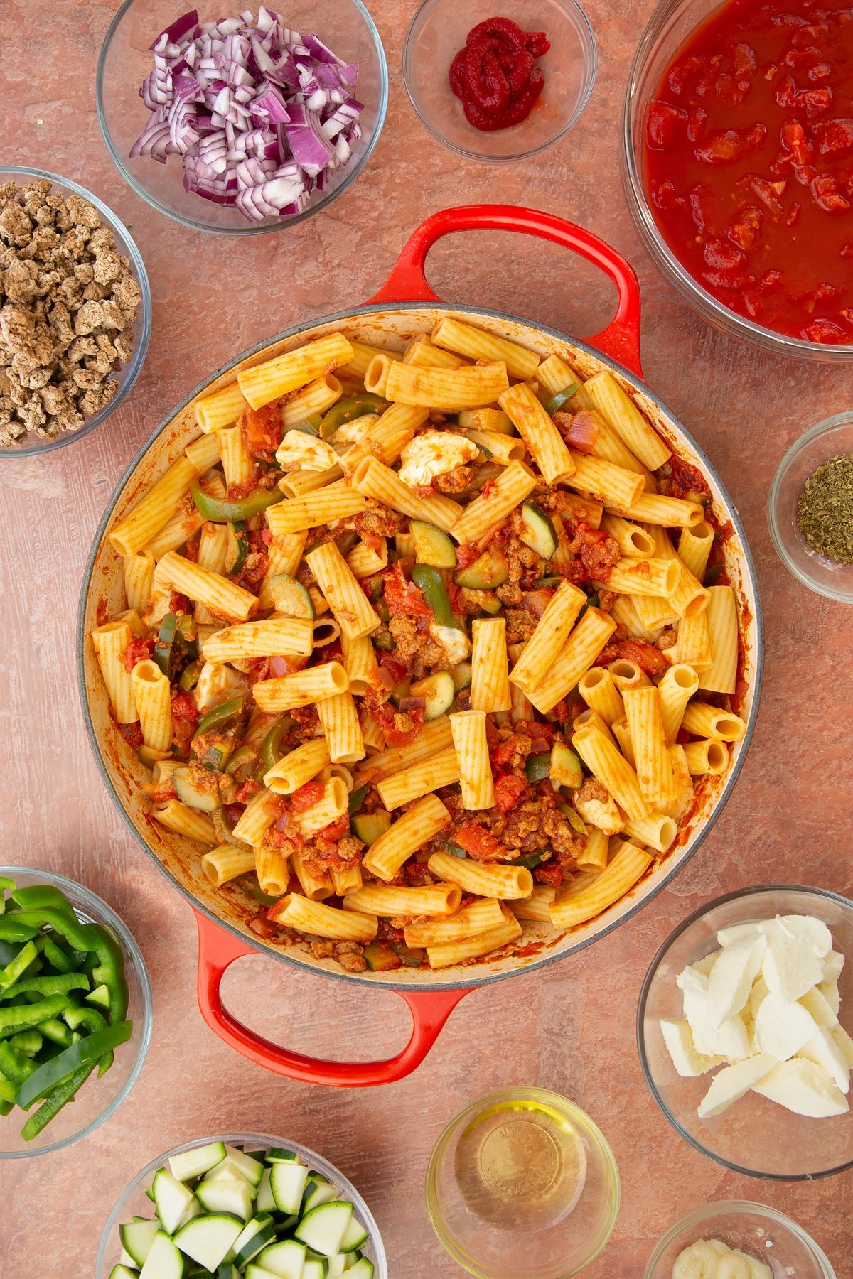 A casserole dish containing a bolognese sauce mixed with cooked pasta and torn mozzarella. Ingredients to make bolognese al forno surround the dish.