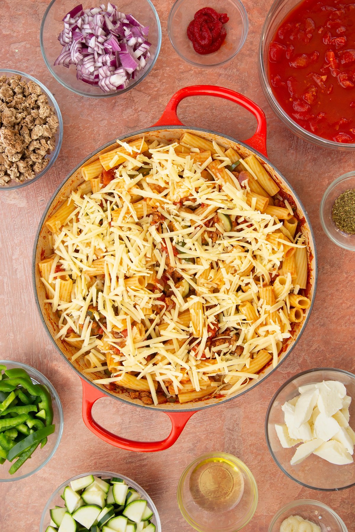 A casserole dish containing a bolognese sauce mixed with cooked pasta and torn mozzarella, topped with grated cheese. Ingredients to make bolognese al forno surround the dish.