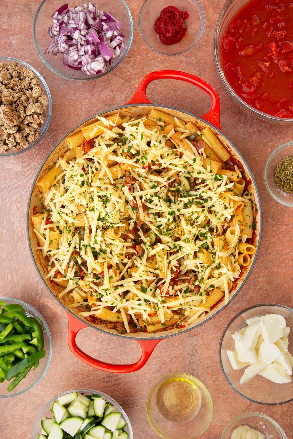 A casserole dish containing a bolognese sauce mixed with cooked pasta and torn mozzarella, topped with grated cheese and chopped parsley. Ingredients to make bolognese al forno surround the dish.