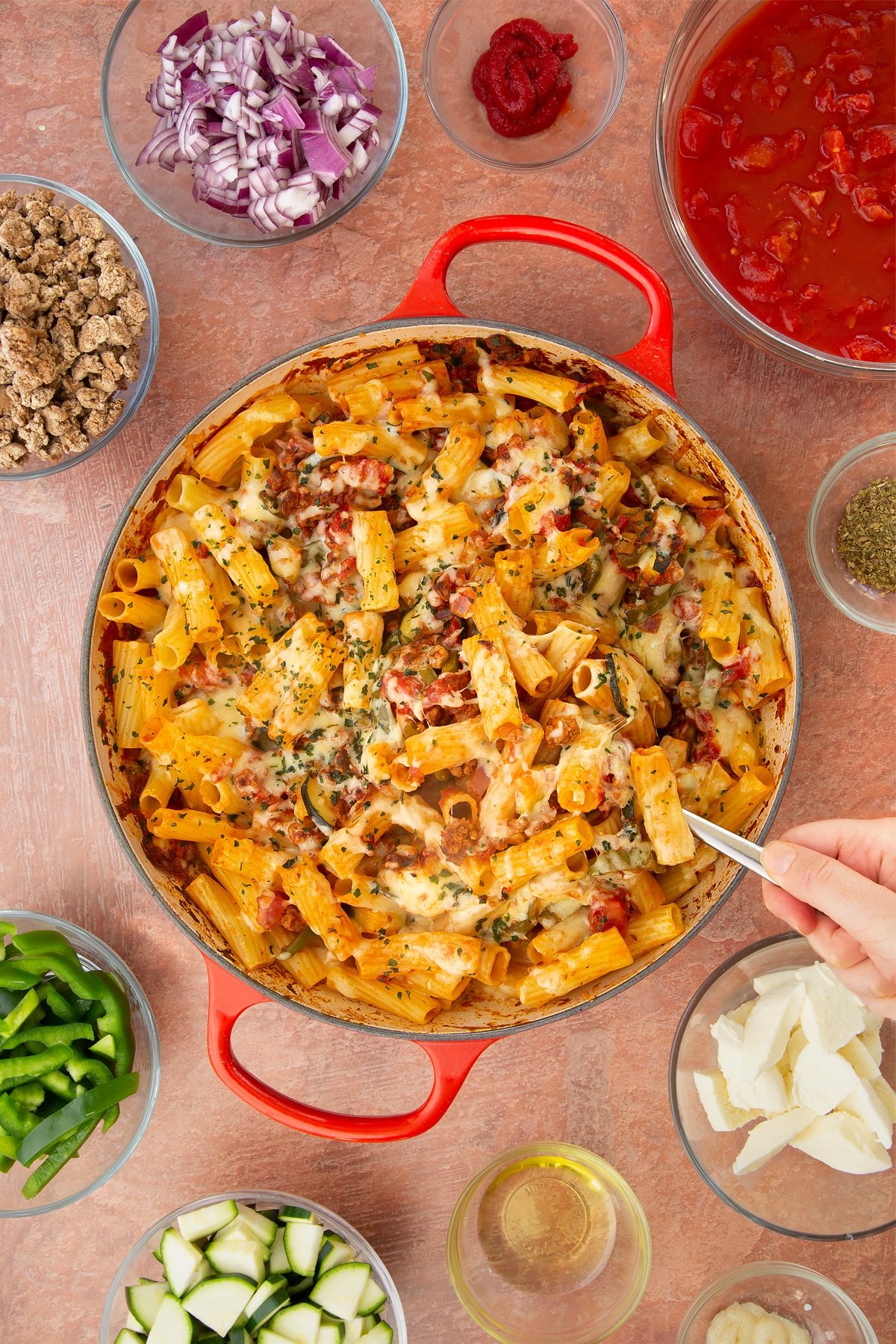 A casserole dish containing freshly baked bolognese al forno. Ingredients surround the dish. A hand holding a large spoon reaches in to serve.