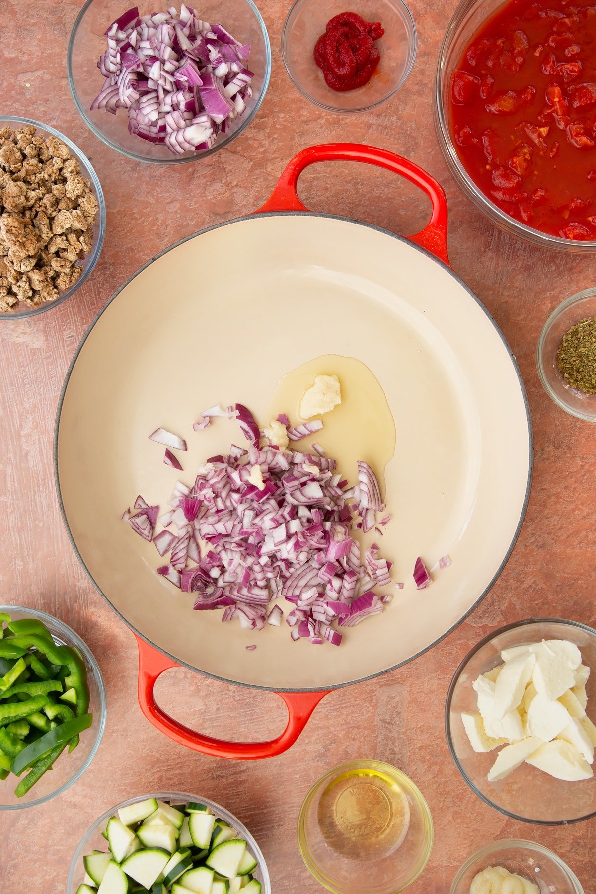 A casserole dish containing oil, chopped red onion and garlic. Ingredients to make bolognese al forno surround the dish.