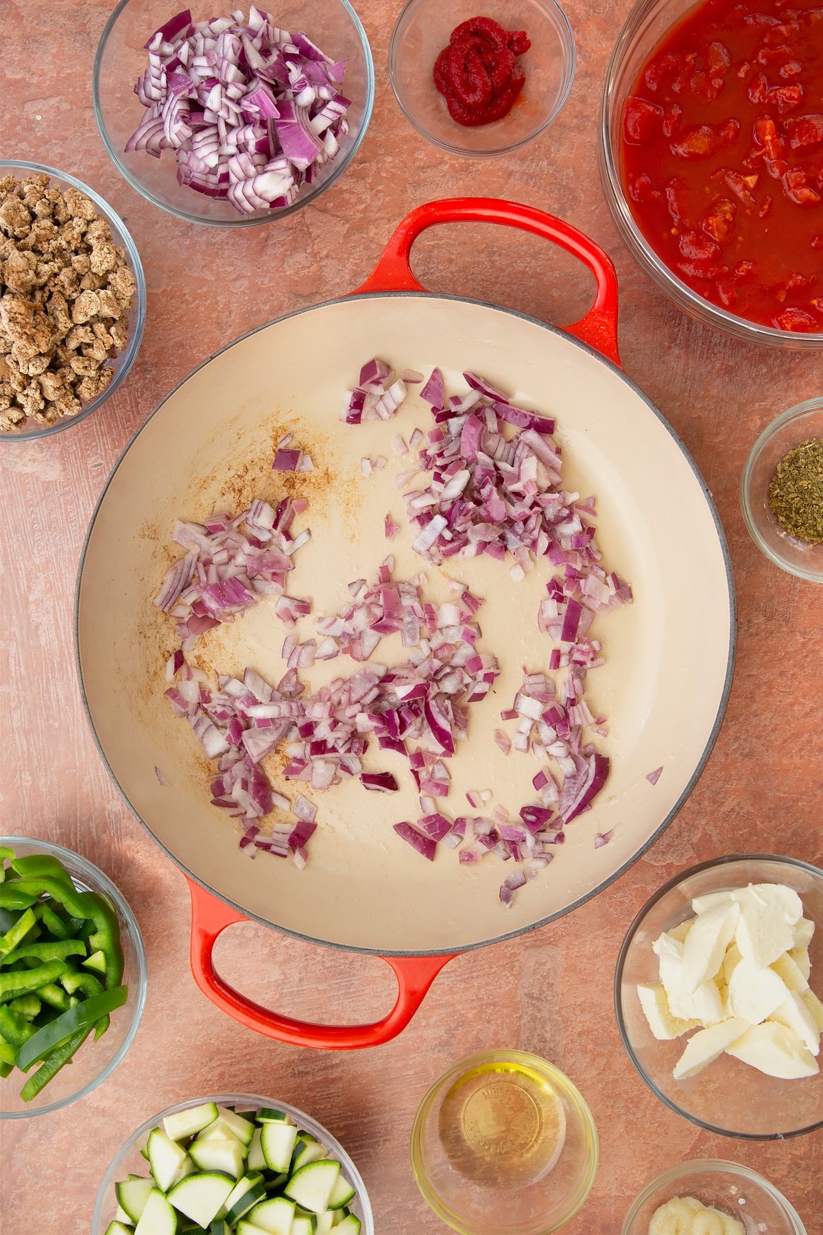 A casserole dish containing fried red onion and garlic. Ingredients to make bolognese al forno surround the dish.