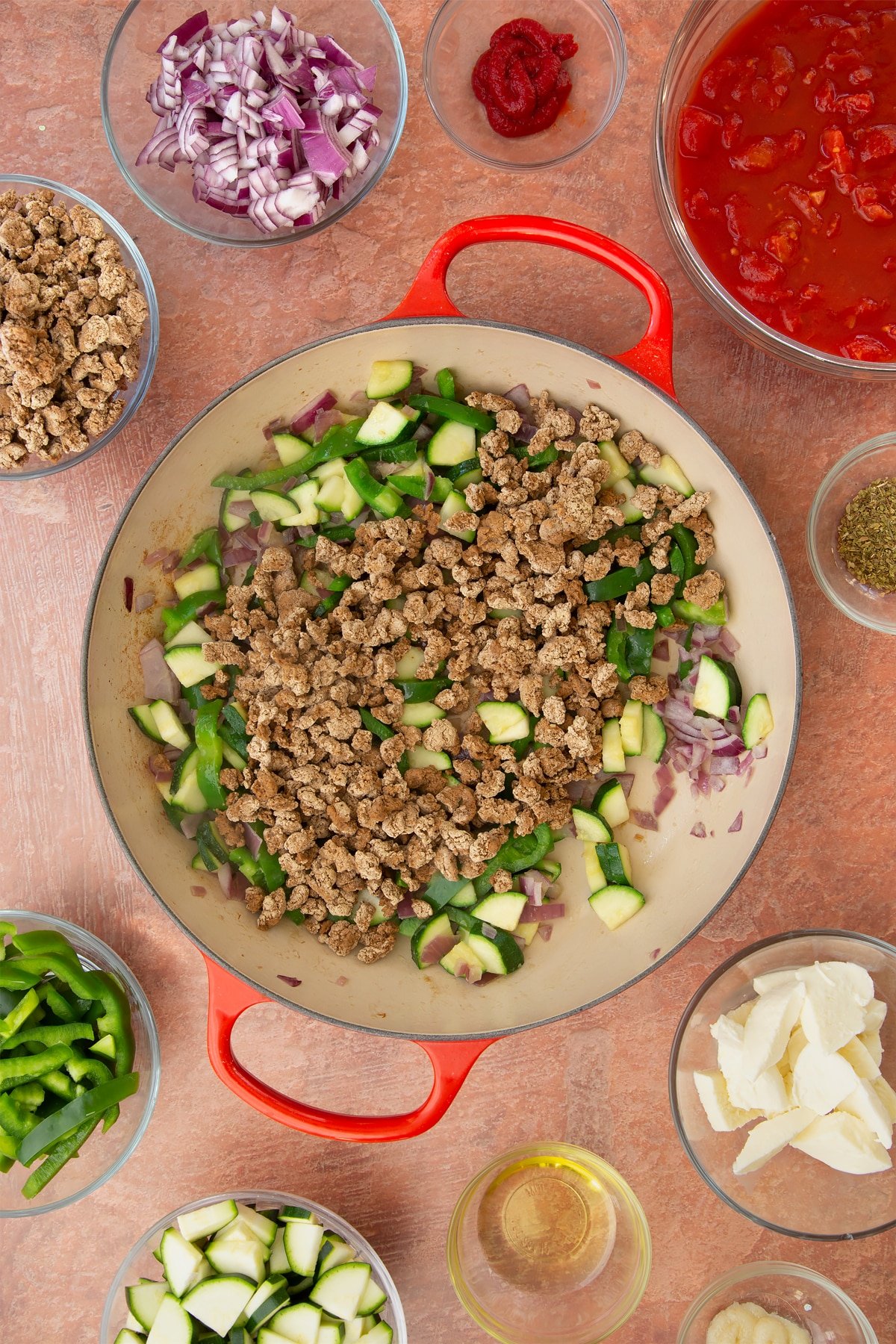 A casserole dish containing fried red onion, garlic, sliced peppers and courgettes. Mince sits on top. Ingredients to make bolognese al forno surround the dish.