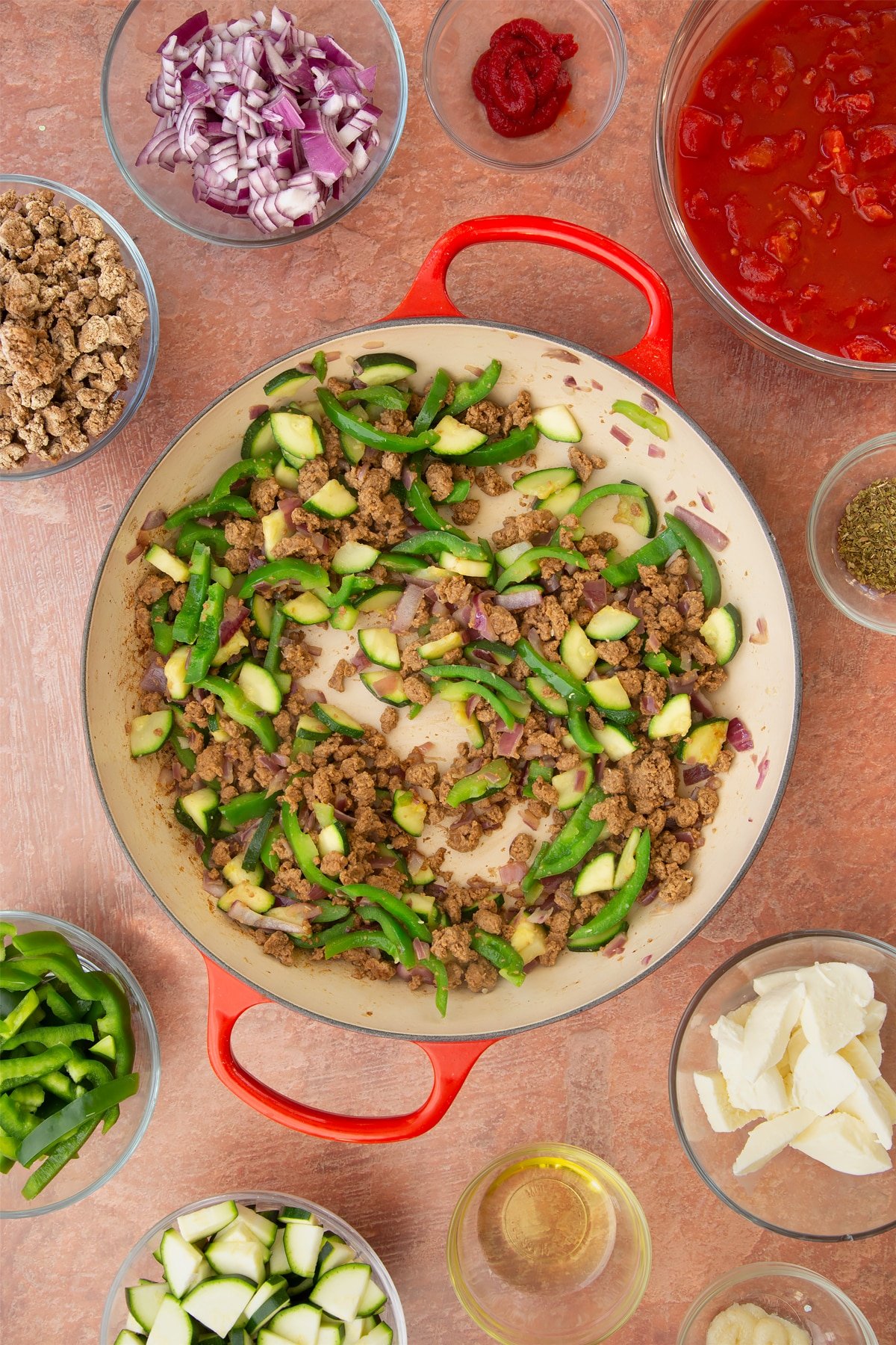 A casserole dish containing fried red onion, garlic, sliced peppers, courgettes and mince. Ingredients to make bolognese al forno surround the dish.