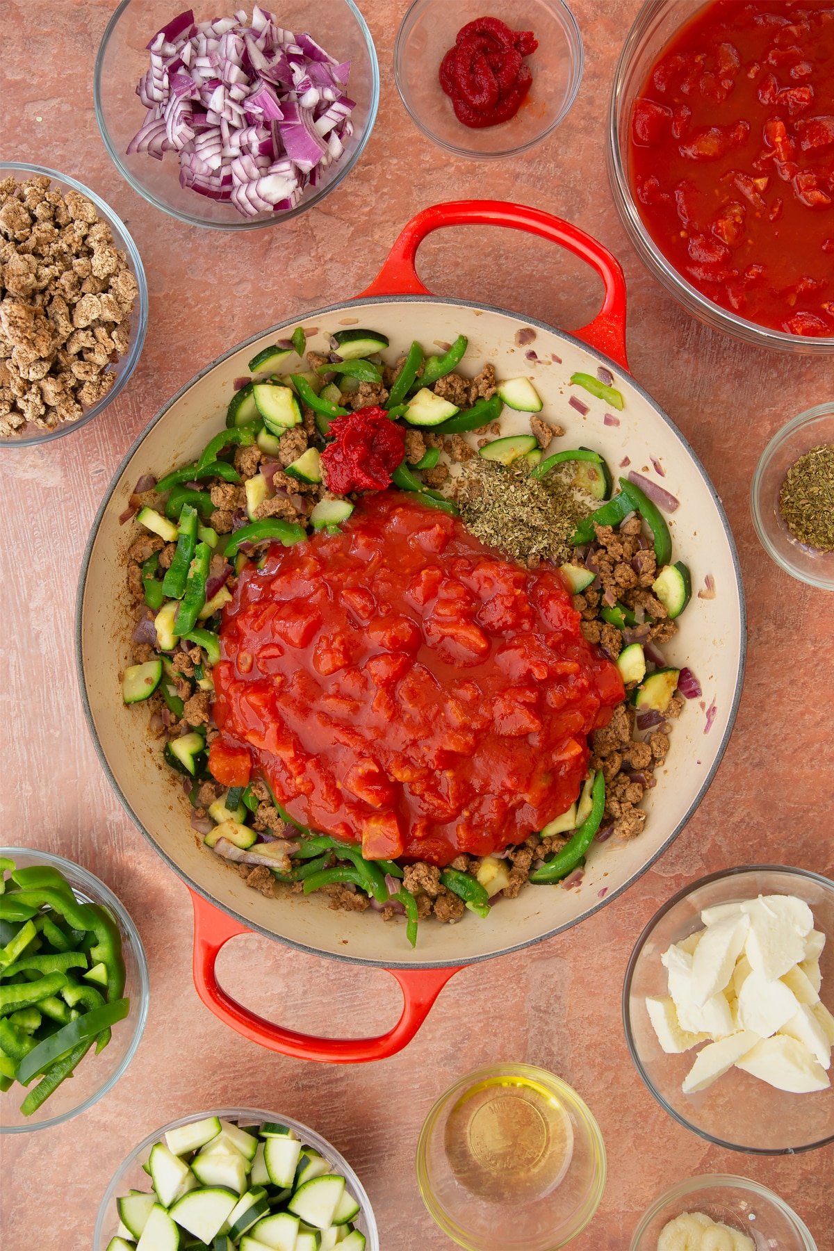 A casserole dish containing fried red onion, garlic, sliced peppers, courgettes and mince. Chopped tomatoes, tomato puree and dried oregano is added. Ingredients to make bolognese al forno surround the dish.