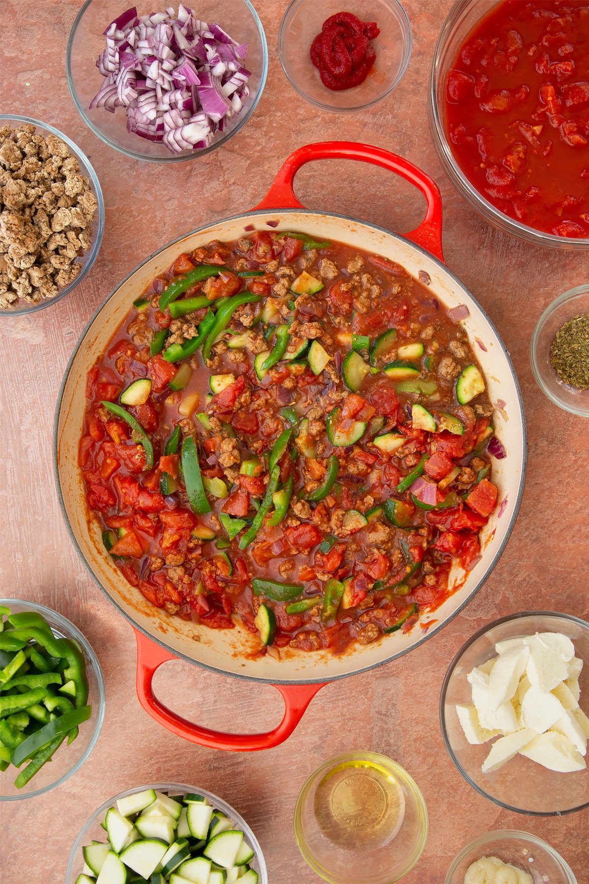 A casserole dish containing fried red onion, garlic, sliced peppers, courgettes, mince, chopped tomatoes, tomato puree and dried oregano. Ingredients to make bolognese al forno surround the dish.